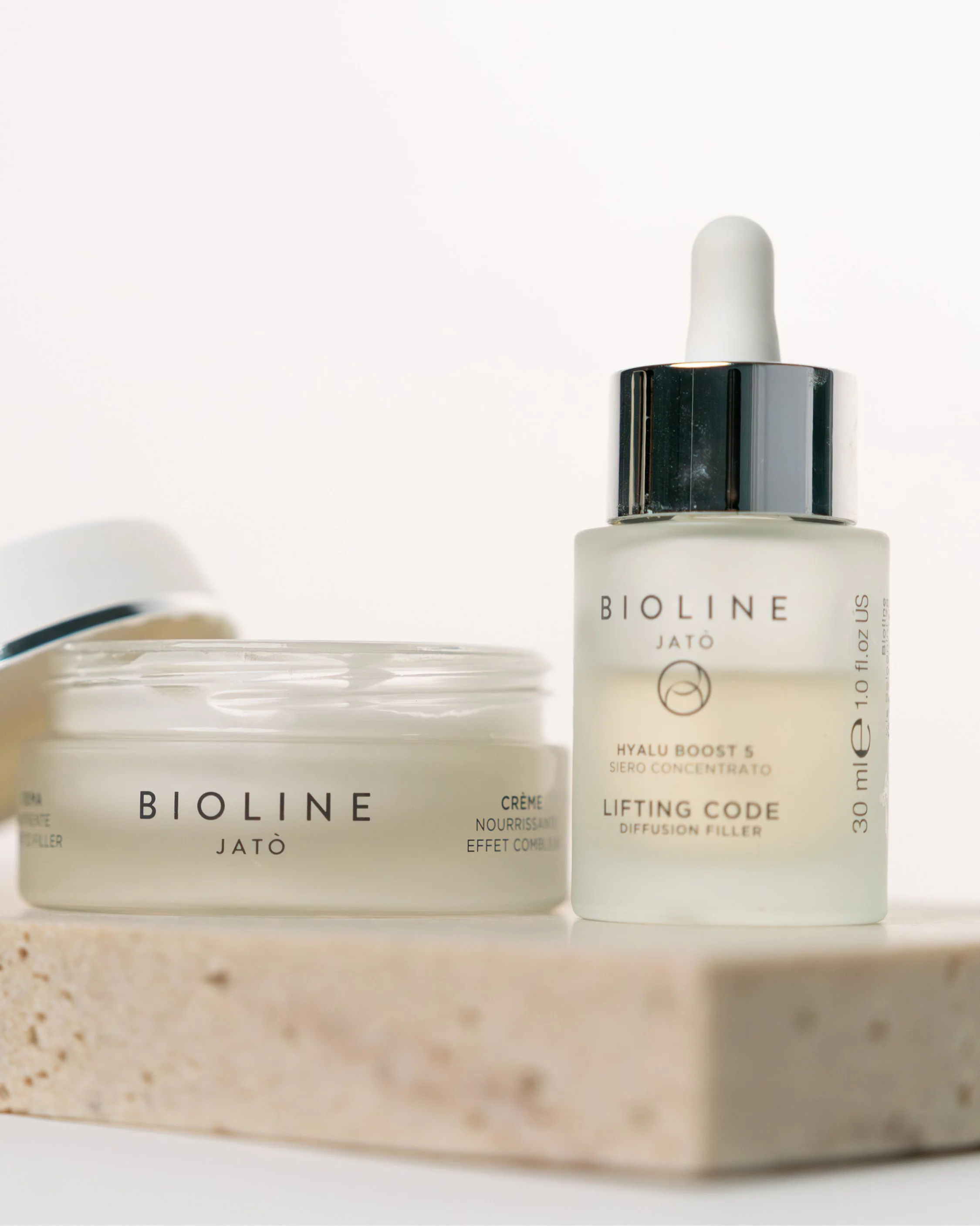 Close-up of skincare products, with a focus on a bottle labeled 'BIOLINE JATÒ Hyalu Boost 5 Lifting Code Diffusion Filler' and a partially visible container behind it, all placed on a beige surface against a plain white background.