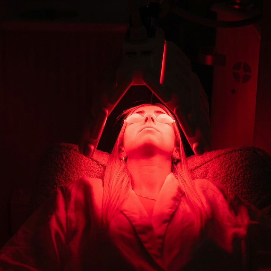 Person lying on a treatment bed receiving red light therapy, with a medical device above their face in a dimly lit room.