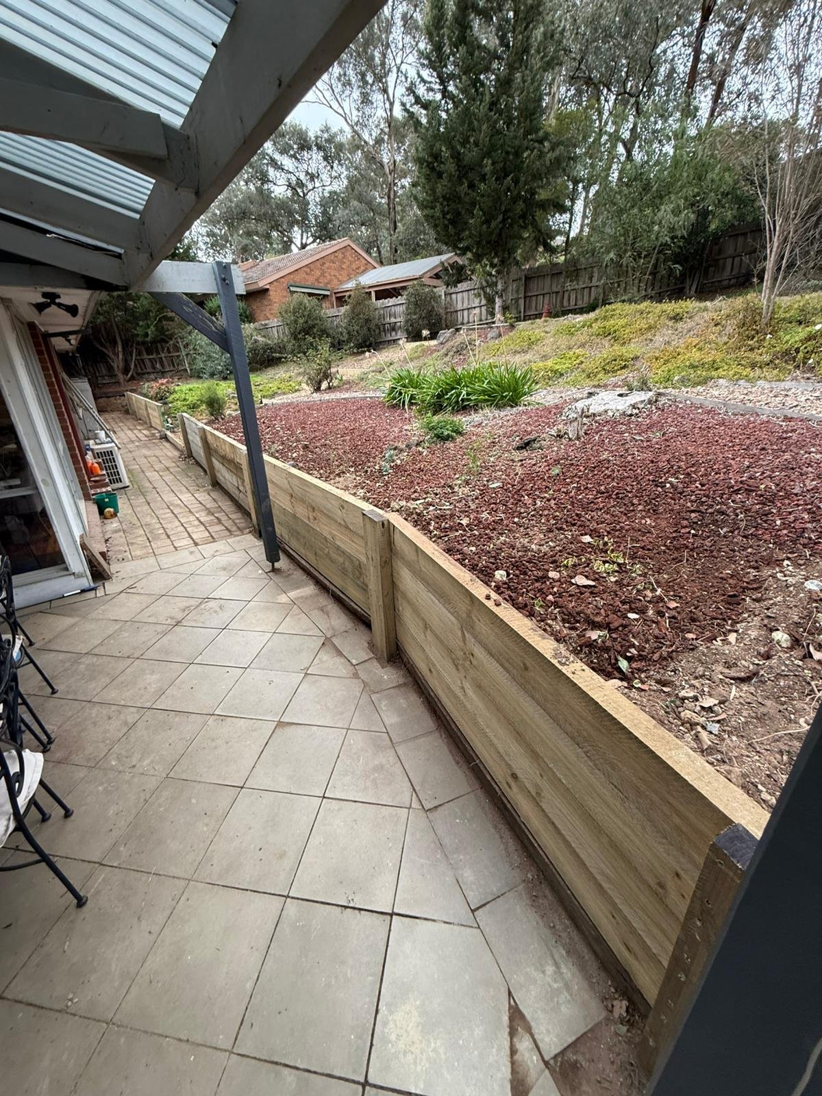 Back patio with tiled and concrete flooring, a wooden retaining wall, and a garden area with mulch, plants, and trees in the background.