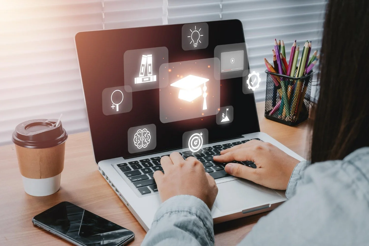 Person using a laptop with holographic icons related to education and knowledge, sitting at a desk with a coffee cup, smartphone, and a container of colored pencils by window blinds.