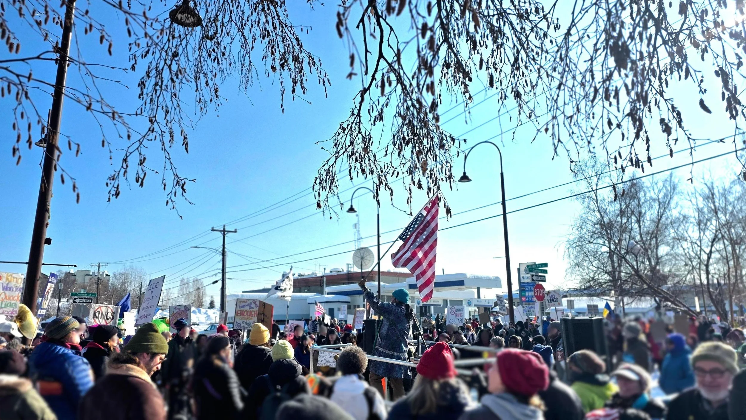 A large crowd gathered outdoors during a rally or protest on a sunny winter day. Many people are wearing hats and warm clothing, holding signs and flags, with one person prominently waving an American flag. The sky is clear and blue, with the tops of