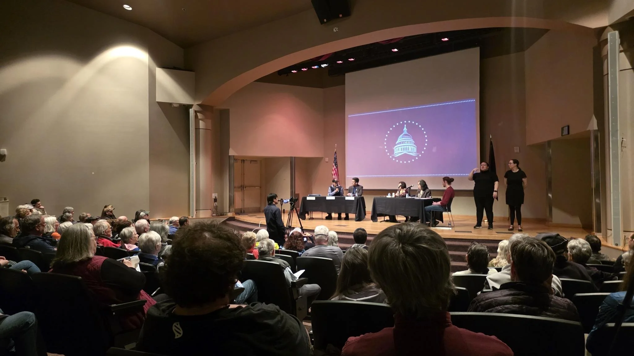 A large group of seated people in an auditorium holding protest signs. The signs read phrases like "Traitor Trump," "Doormat Dan," "No Kings," and "Chicken Nick." A microphone stand is set up at the front of the room.