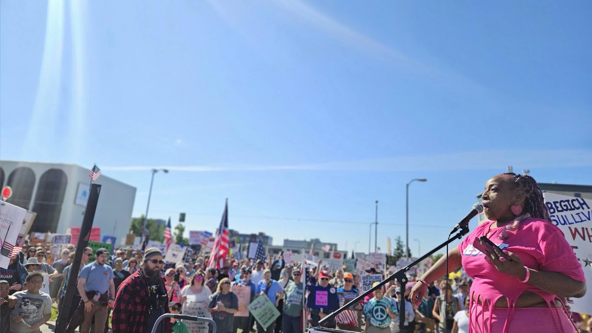 A woman in a pink outfit speaking into a microphone at a protest or rally, with a large crowd holding signs in the background.