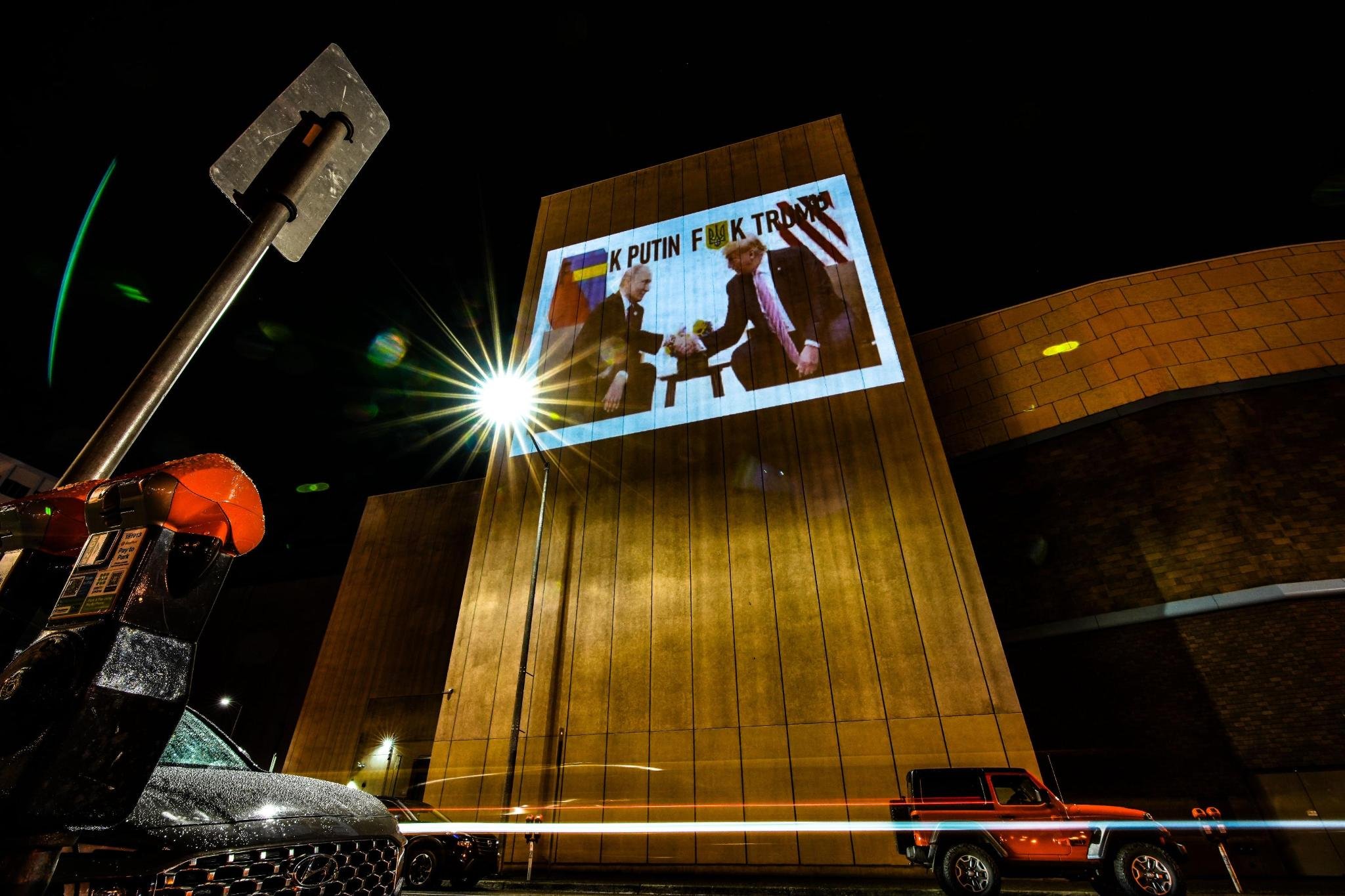 Nighttime street scene with a large building and streetlight. A digital billboard shows a photo of two men shaking hands, with the words 'K PUTIN F**K TRUMP' visible. Cars parked along the street, including an orange vehicle in the foreground.