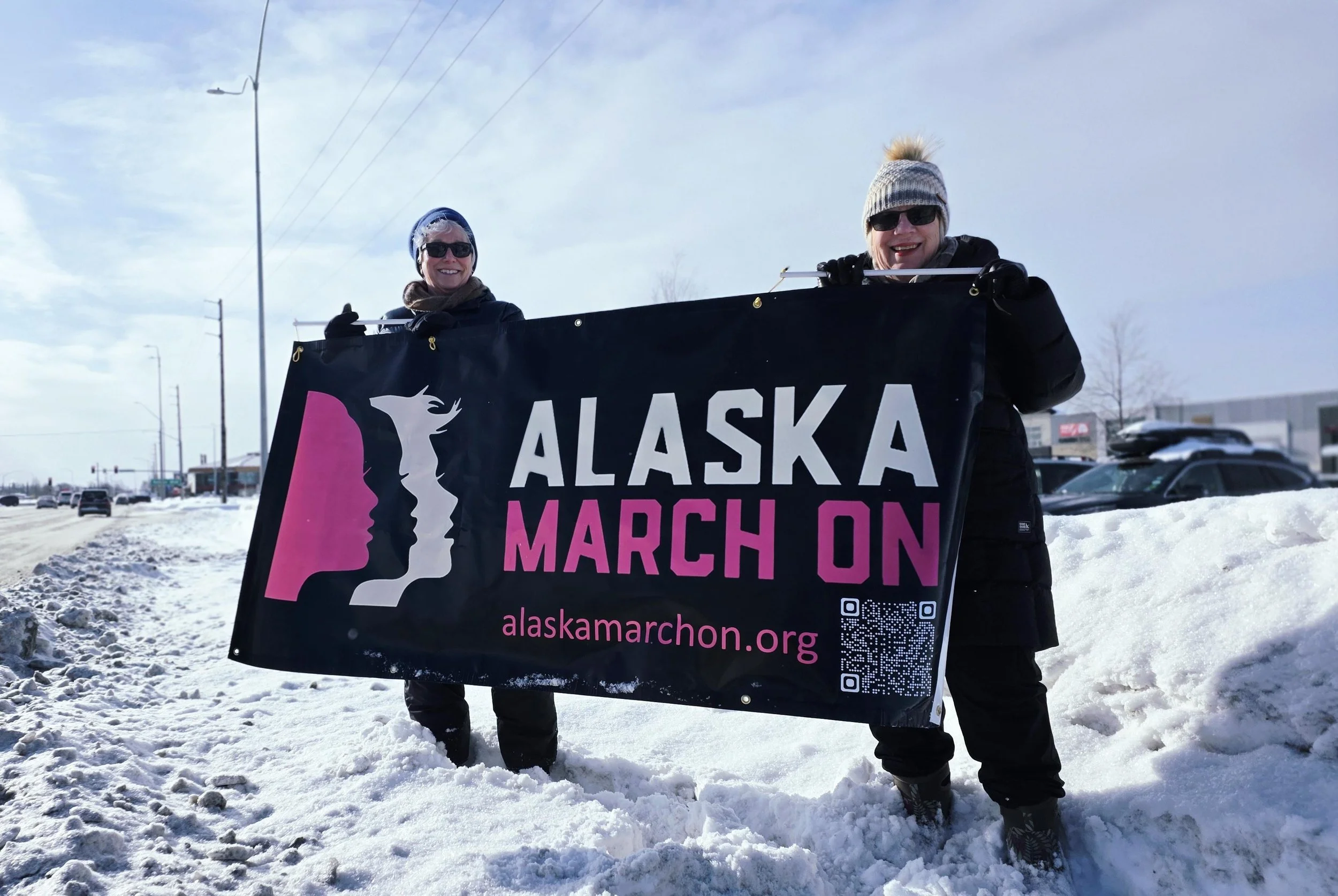 Two women standing in snow holding a black banner that says "Alaska March On" with a stylized pink and white logo and a website URL "alaskamarchon.org".