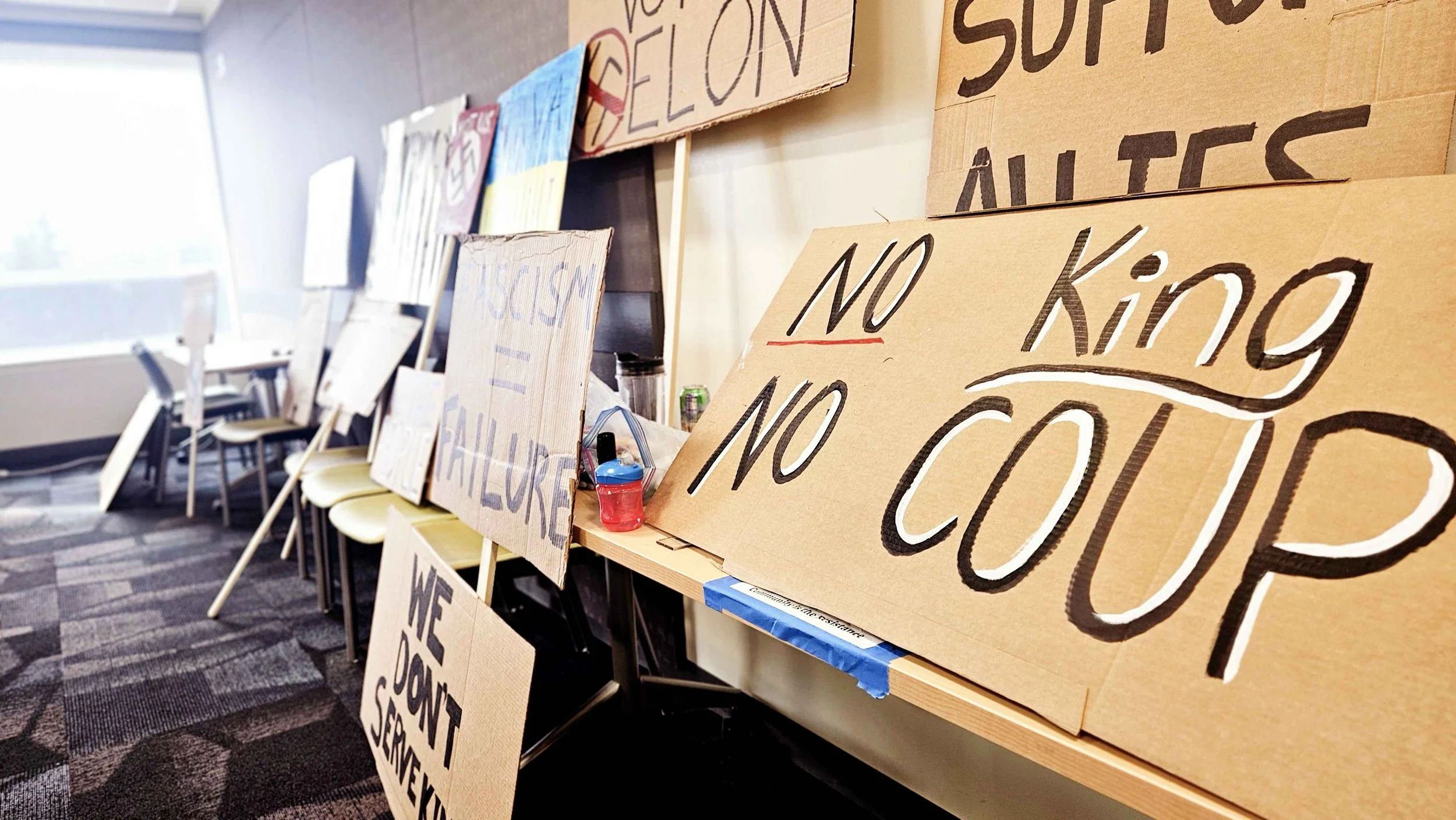 A row of cardboard protest signs leaning against a wall and chairs, displaying messages like "No King" and "We Don't Serve" in an office or conference room with a window in the background.