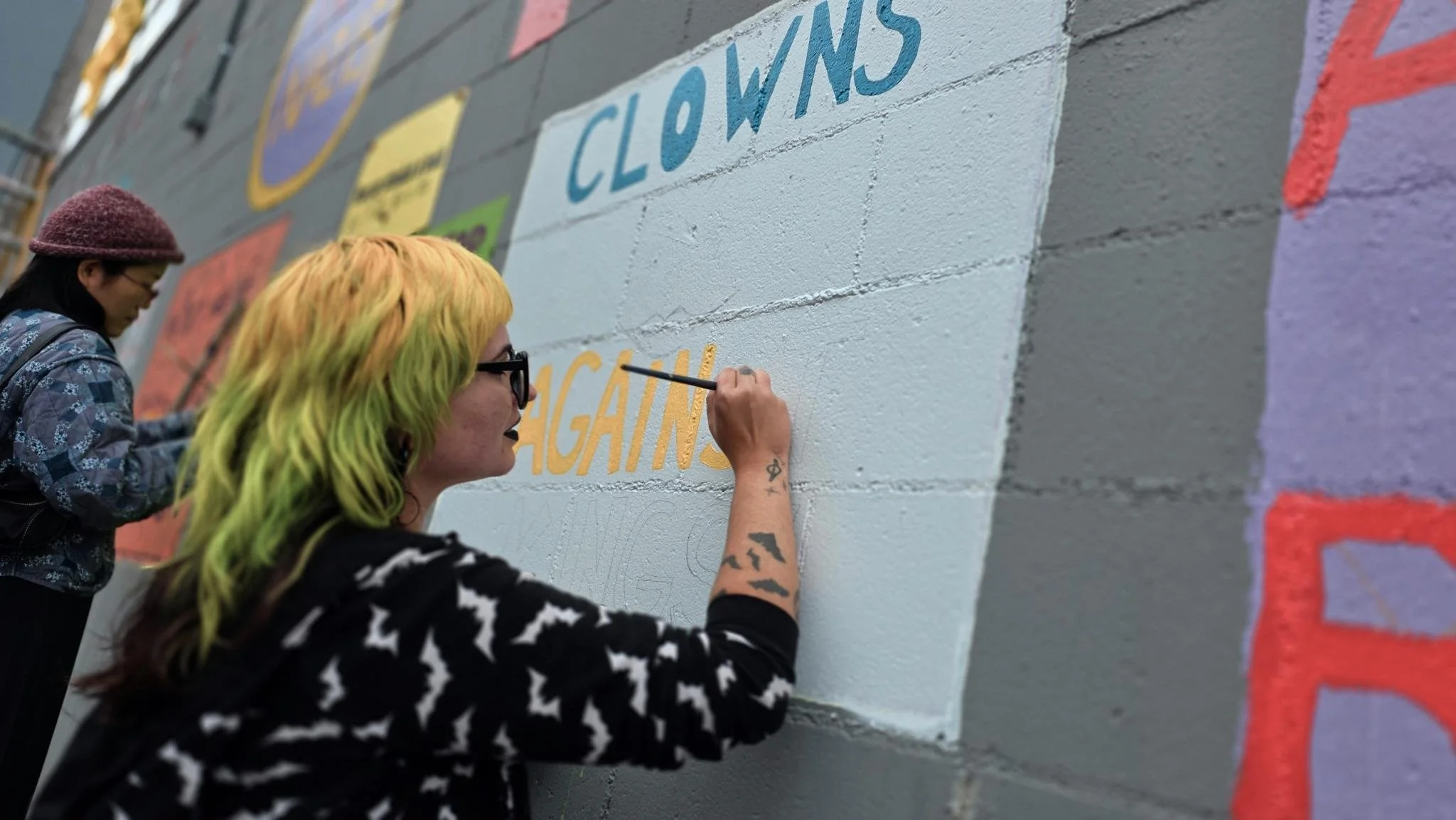 Two women painting a mural on a gray brick wall, with one woman having yellow and green hair and the other woman wearing glasses and a purple hat.