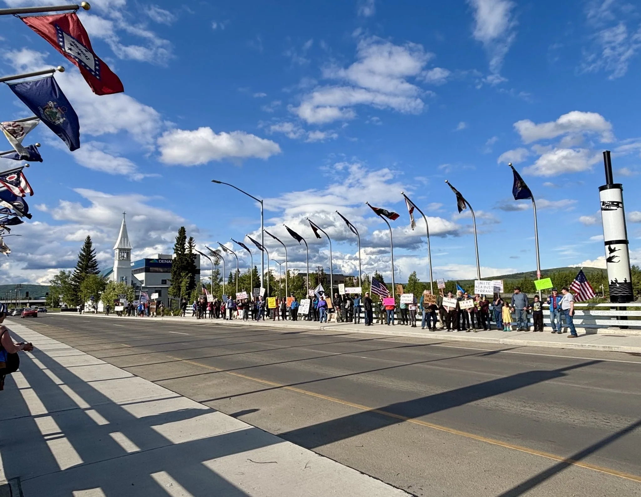Crowd of people protesting on a sidewalk with signs and American flags, with flags flying on poles and a church with a steeple in the background.