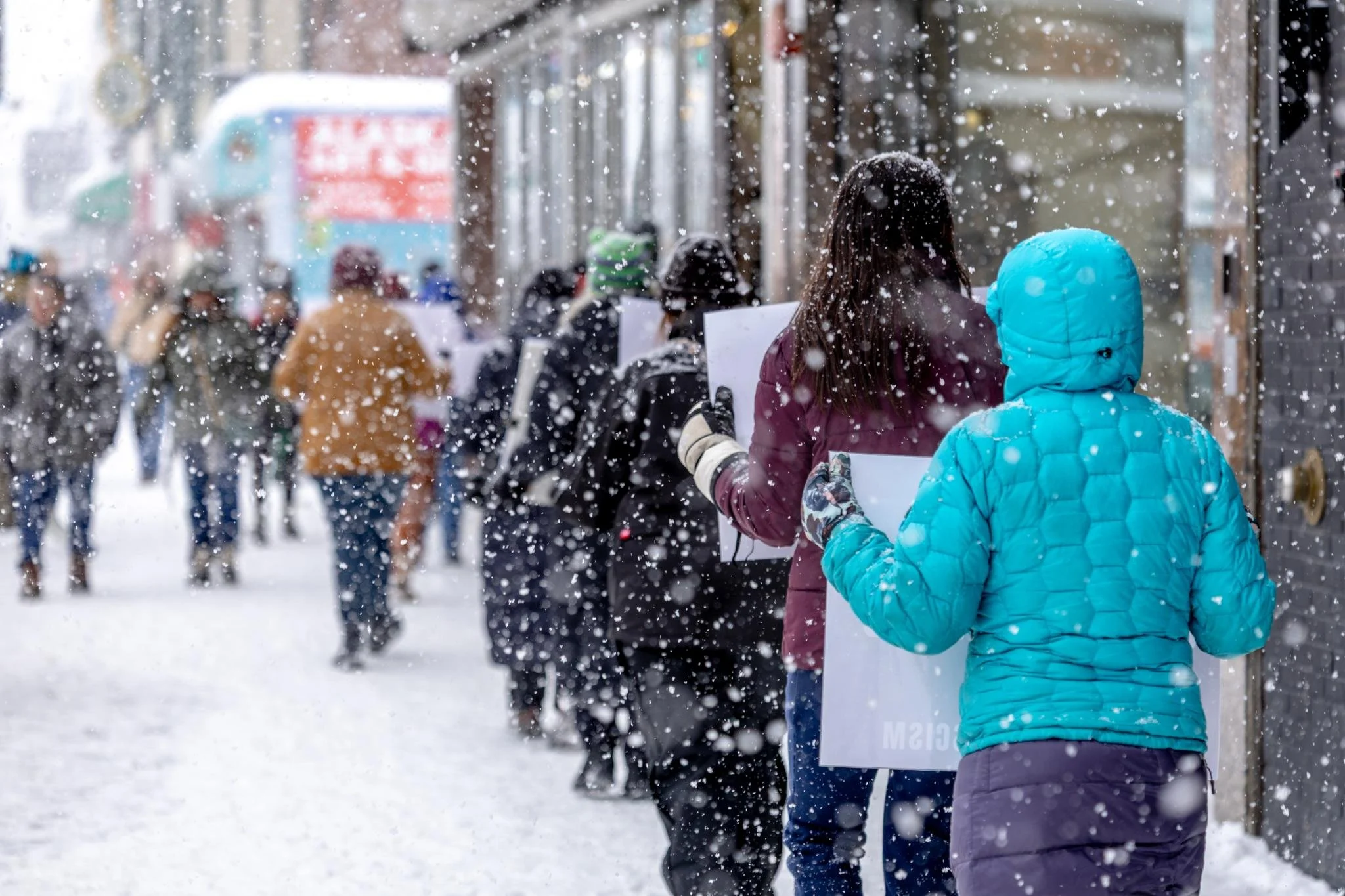 A crowd of people are standing in line outside on a snowy city street, wearing winter clothing and holding signs.