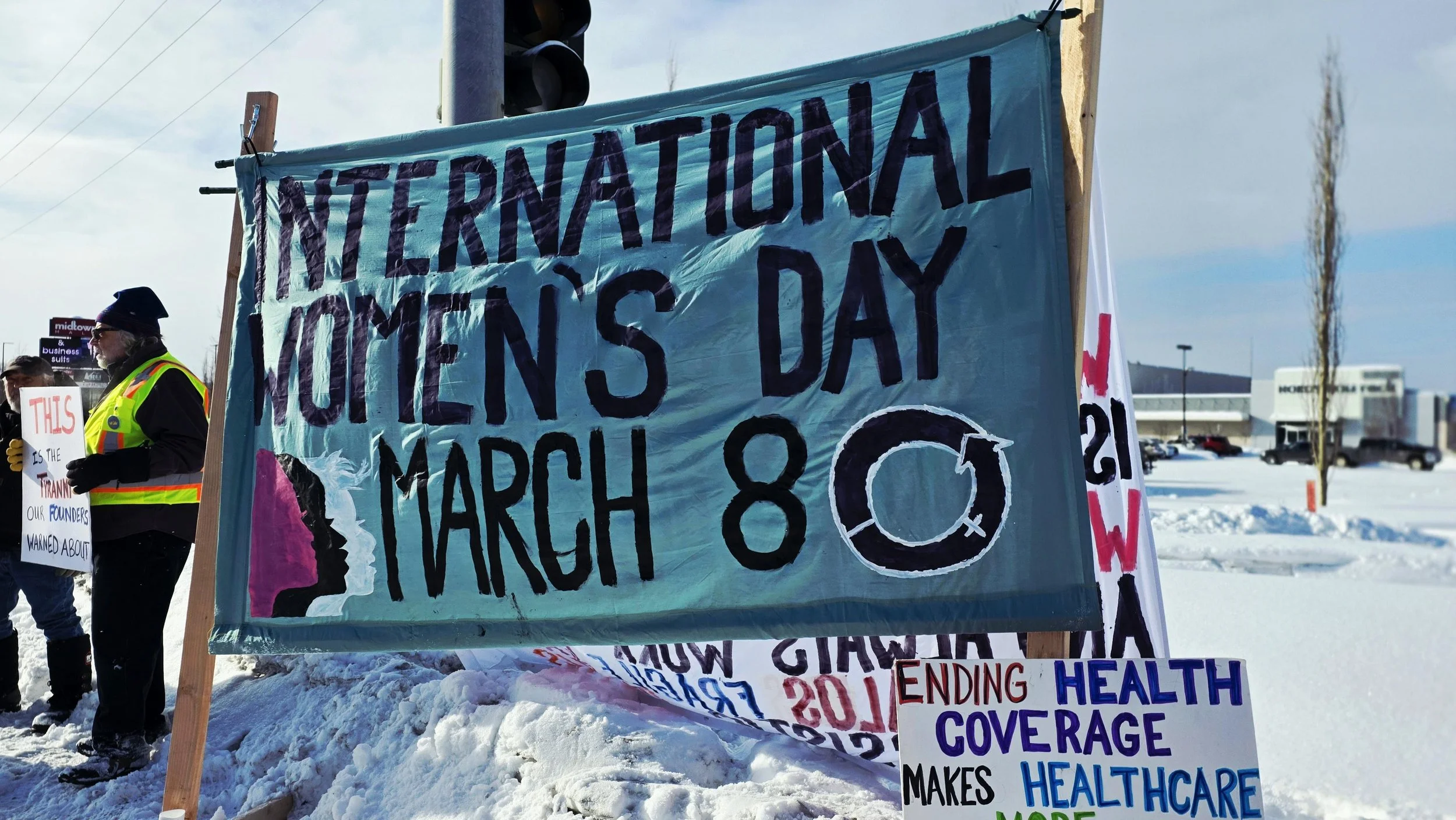 A protest with people holding signs and banners for International Women's Day on March 8th, during winter with snow on the ground.