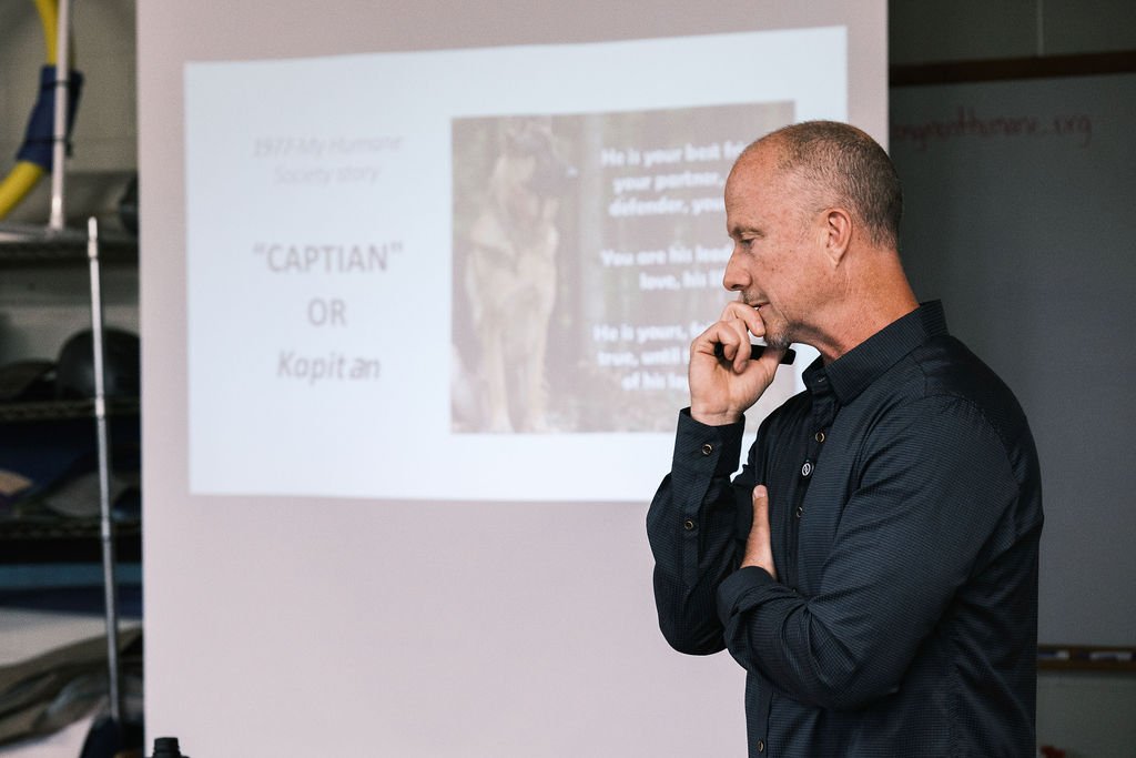 A man standing in front of a presentation slide, thoughtfully touching his chin, with a finger on his lips, and looking at the screen.