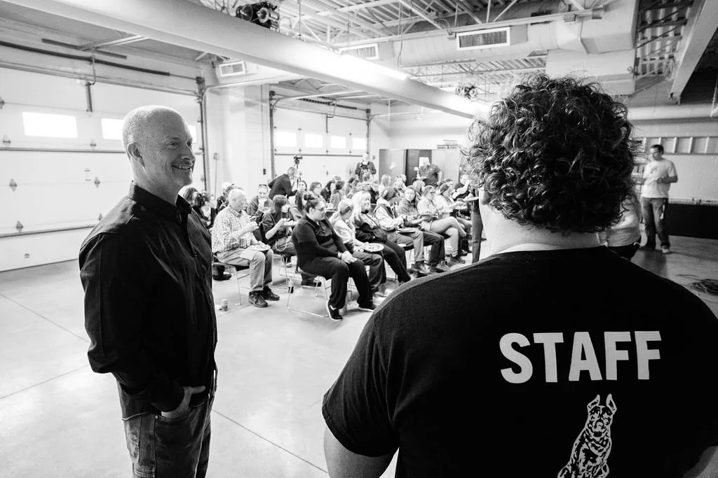 People gathered in a garage-like space, with two individuals in the foreground, one smiling and the other wearing a 'STAFF' t-shirt, speaking to a seated audience.
