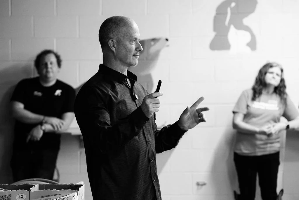 A man giving a presentation, gesturing with his hands, with two women standing behind him against a white brick wall.