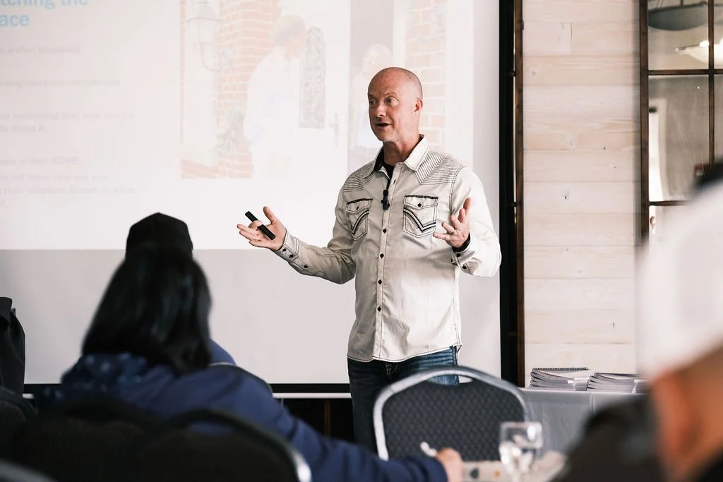 A man giving a presentation to an audience in a room with a wooden wall and a window, with a projection screen behind him