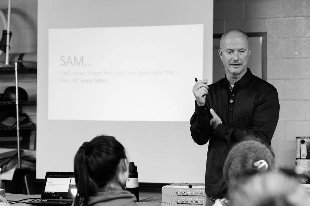 A man giving a presentation to a group of people in a classroom or meeting room, with a slide displayed on a projection screen in the background.