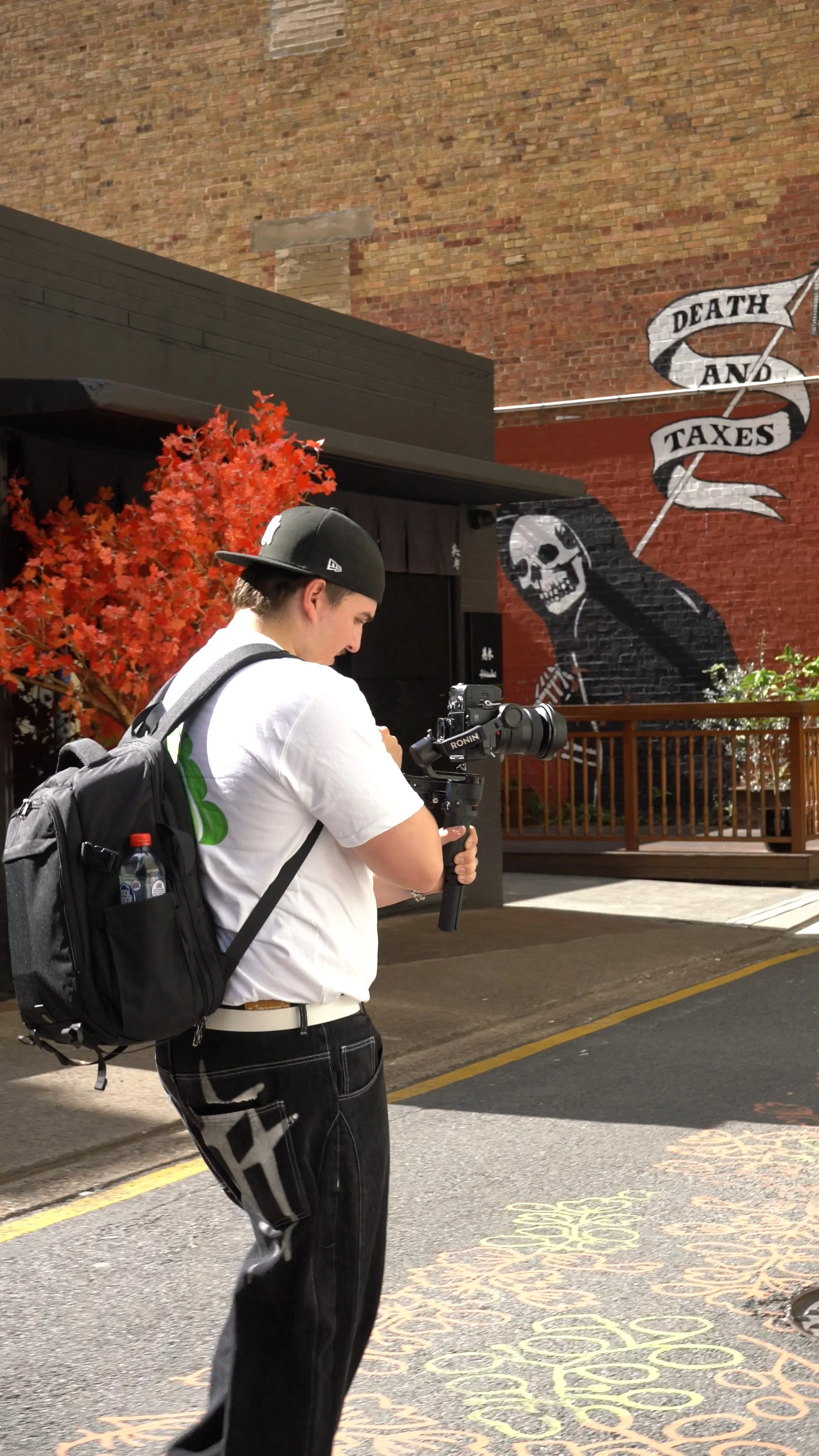 A young man with a backpack and baseball cap filming with a camera on a street, with a mural of a death figure holding a scythe and a banner reading 'Death and Taxes' in the background.