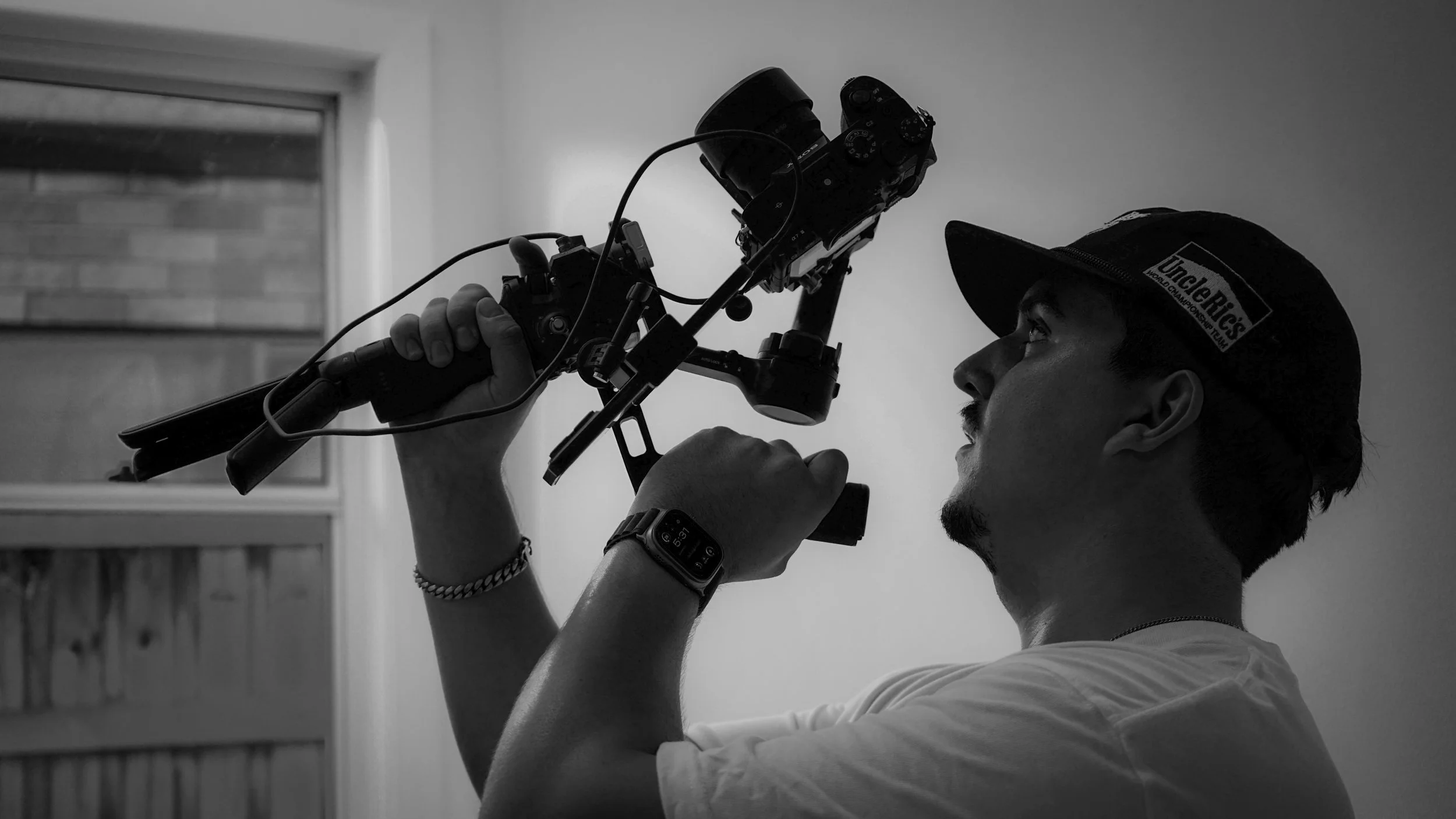 A man wearing a hat and a smartwatch is holding a professional camera on a stabilizer, preparing to take a photograph indoors, with a window in the background.