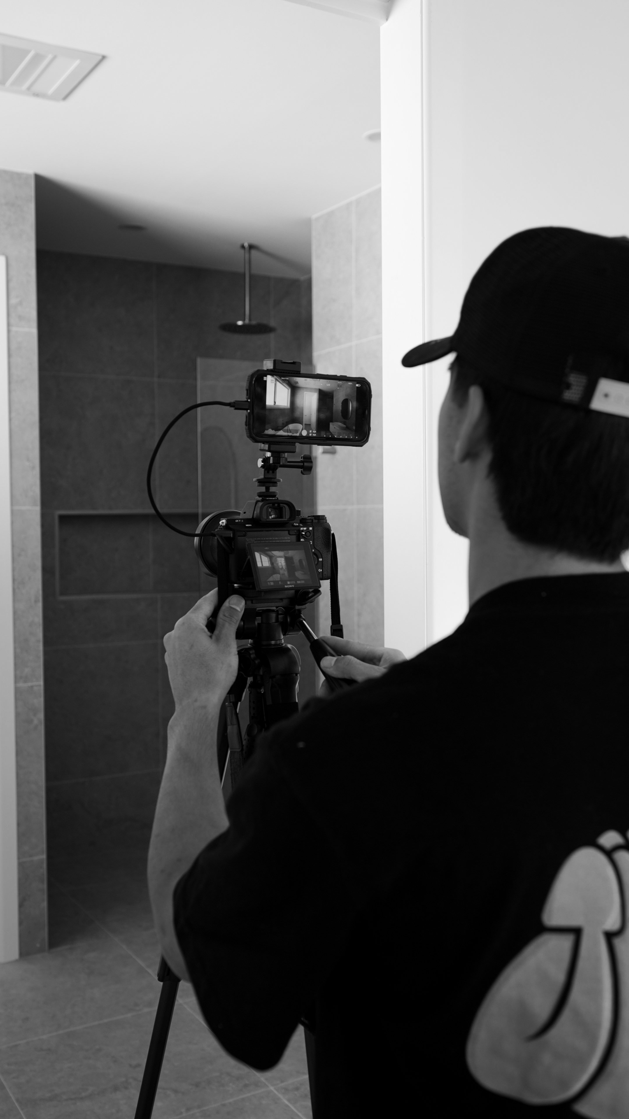 A person operating a camera on a tripod, filming in a bathroom with a shower in the background.