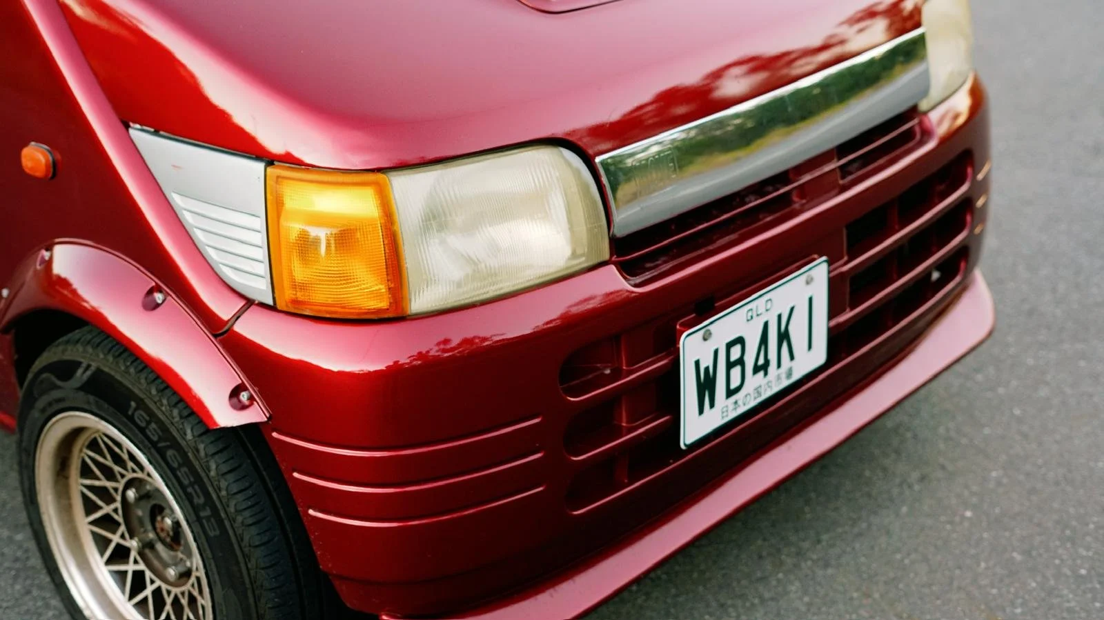 Close-up of the front of a red vintage car with an orange and clear headlight, a Mitsubishi-style front grille, and a California license plate reading 'WB4K1'.