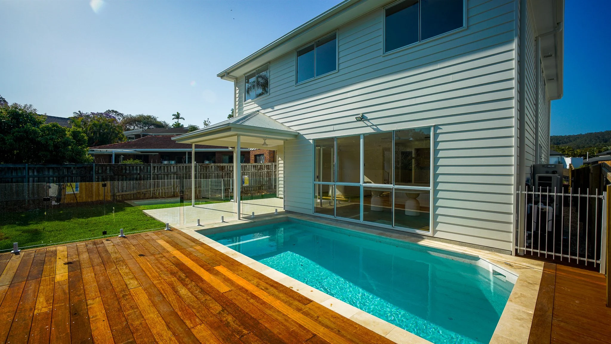 Backyard of a house with a small swimming pool, wooden deck, glass safety fence, large windows, and a porch with a ceiling fan, with neighboring houses and trees in the background.