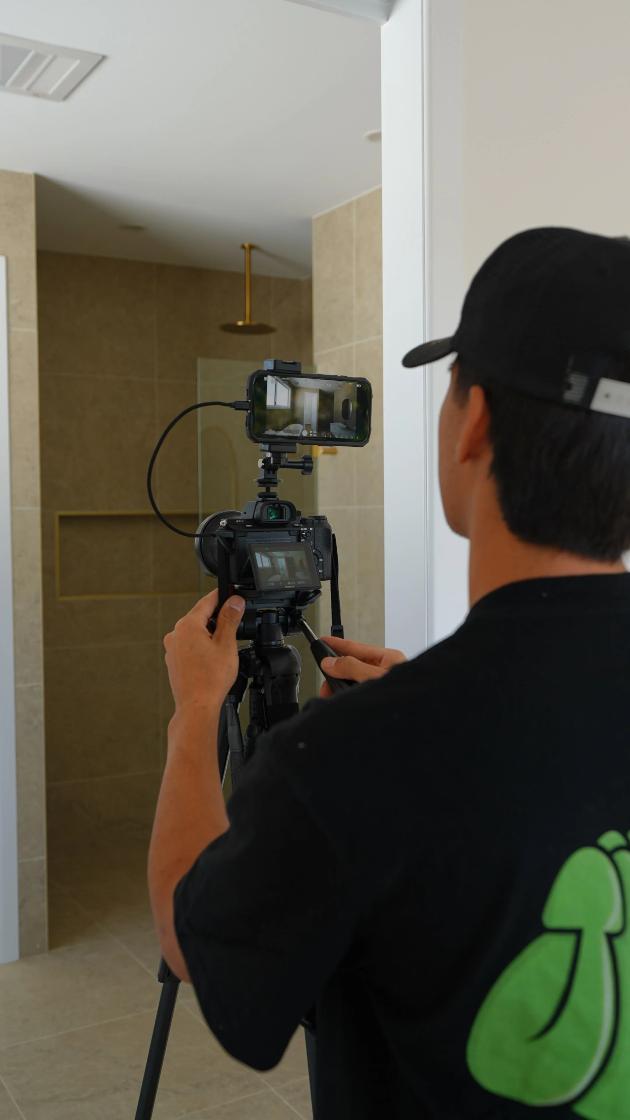 A person wearing a black cap and black T-shirt operating a professional camera on a tripod, filming or photographing the bathroom shower area.