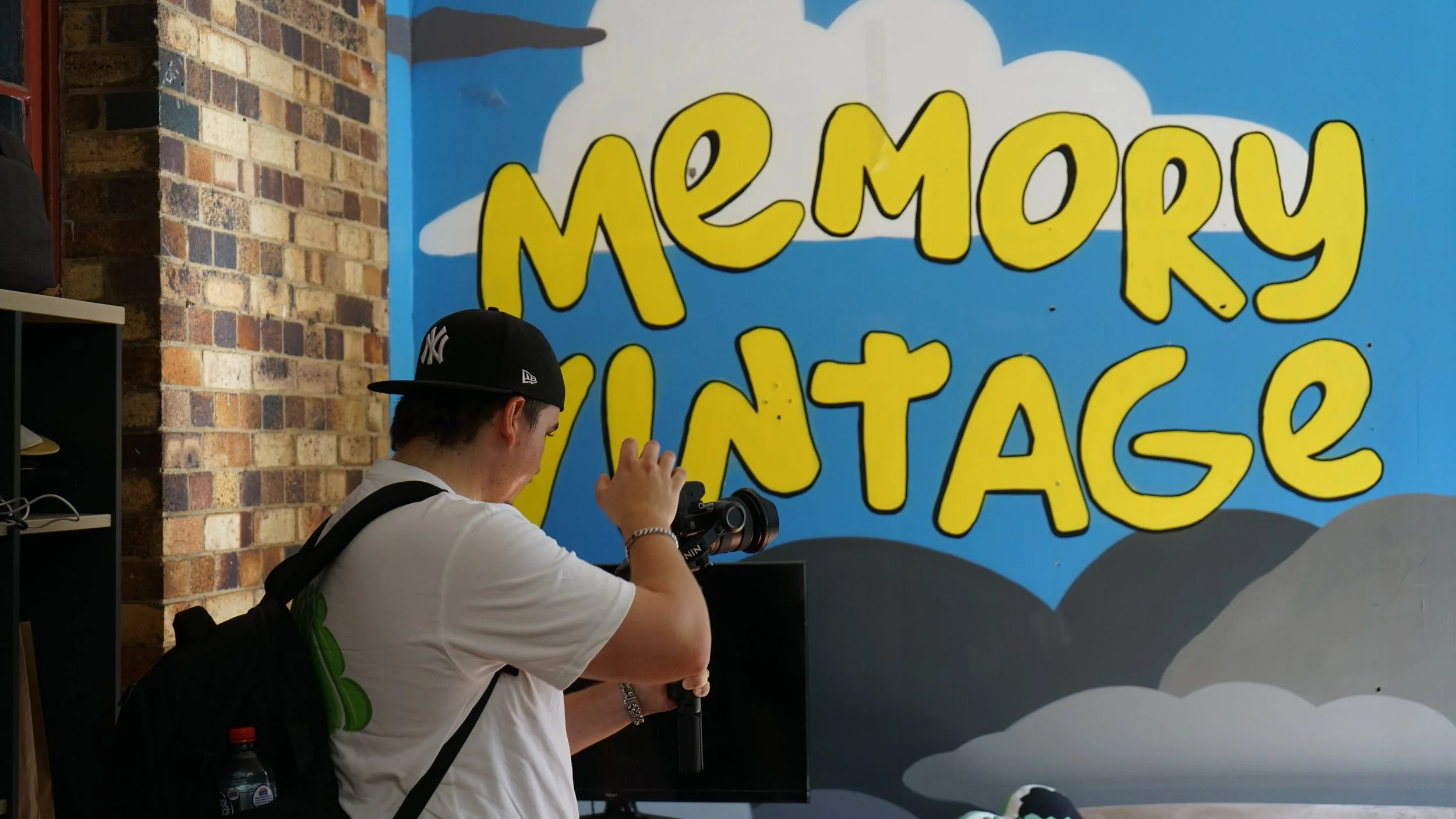 A young man with a black backpack, white shirt, and black cap taking a photo with a camera in front of a wall with colorful graffiti that reads 'Memory Vintage' on a blue cloud background.