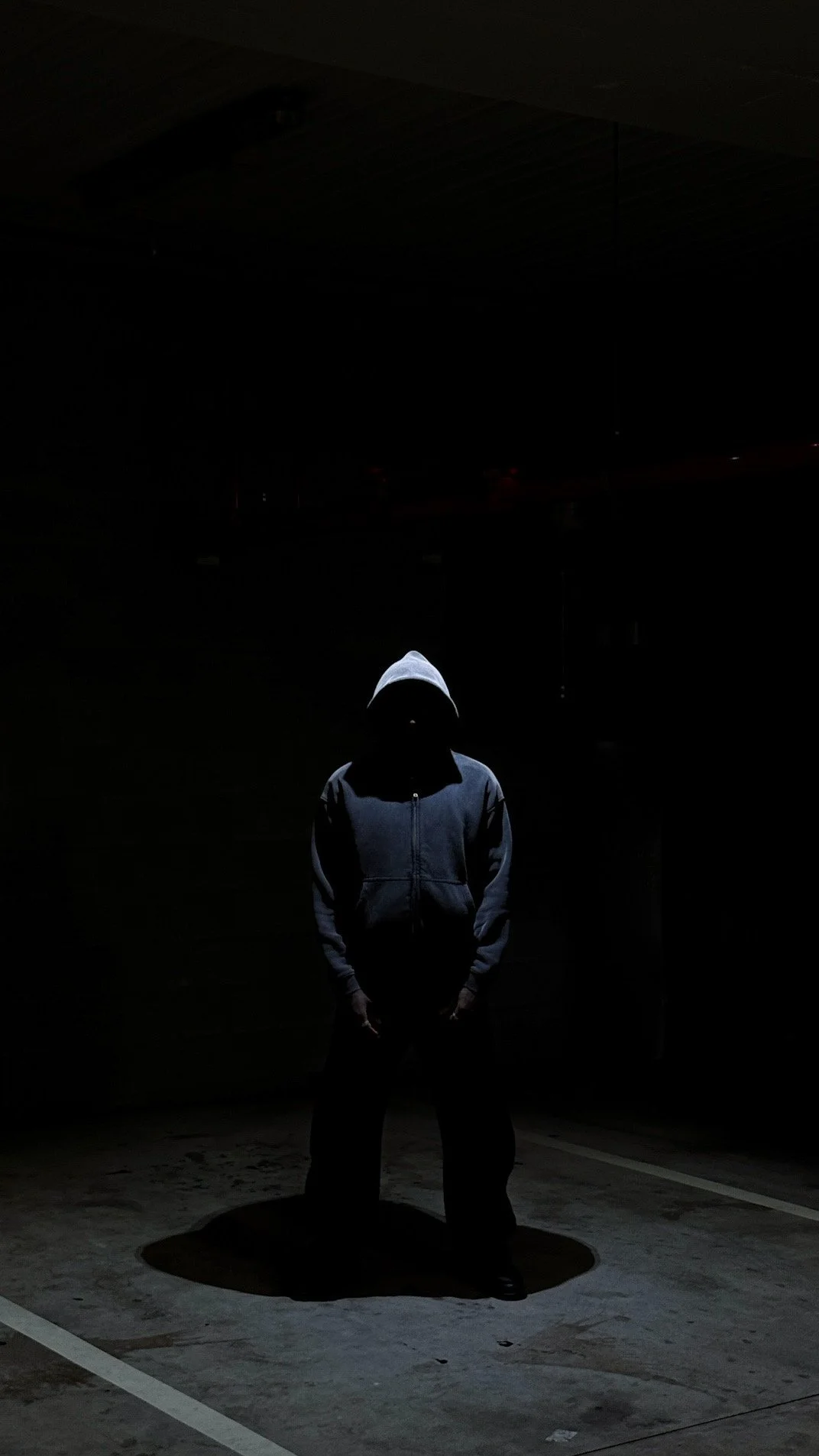 Person wearing a hoodie standing in darkness, illuminated by a faint light, casting a shadow on the ground.