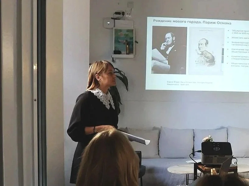 A woman giving a presentation in front of a screen with a slide about Paris Ohsman and her birth of a new city. The room has a couch with pillows, a small table, and some projector equipment.