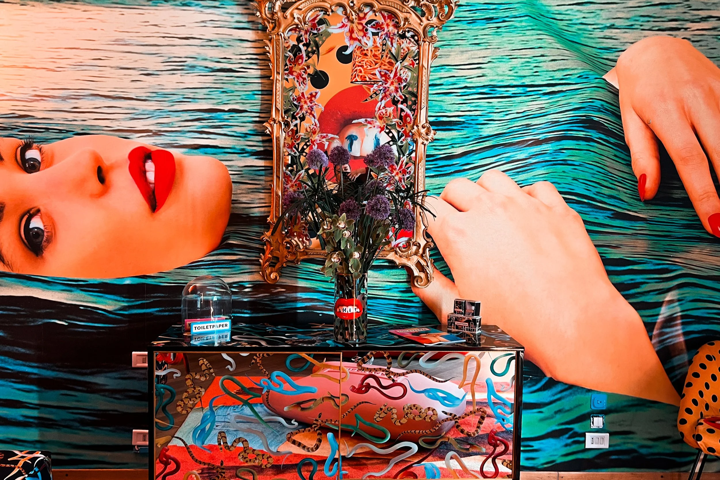 Colorful art installation with a surreal depiction of a woman's face and hands, a mirror with floral and snake decorations, purple flowers in a vase, and various objects including a small sign labeled 'TOILETPAPER', with a vibrant water-themed background.