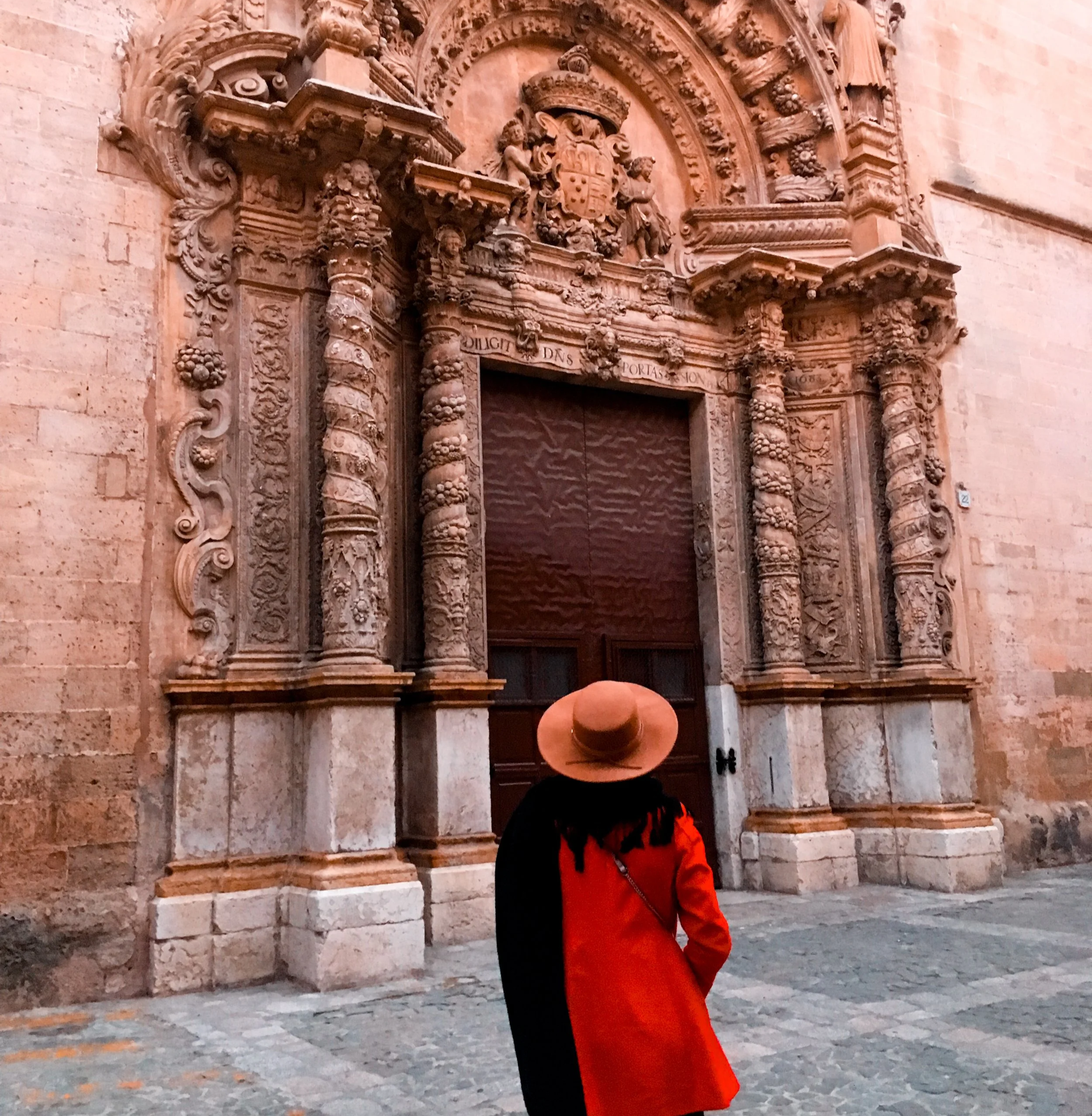 A person wearing a red jacket and a tan wide-brimmed hat standing outdoors in front of an ornate stone archway with intricate carvings and a carved coat of arms. The person has their head tilted down and is facing the archway.