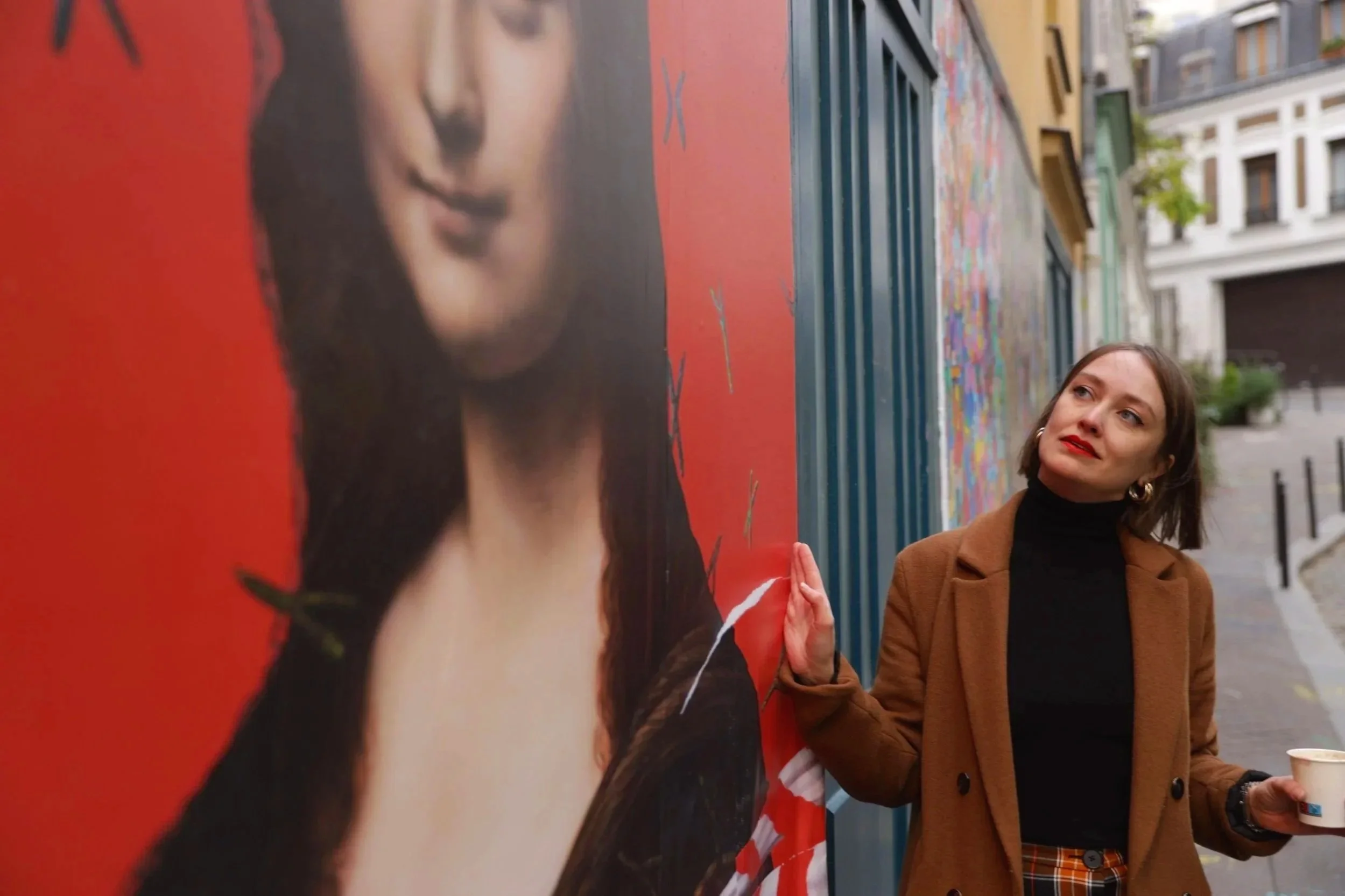 A woman with short brown hair, wearing a brown coat, black turtleneck, and plaid pants, standing outdoors, holding a cup, posing next to a large billboard with a woman's face.