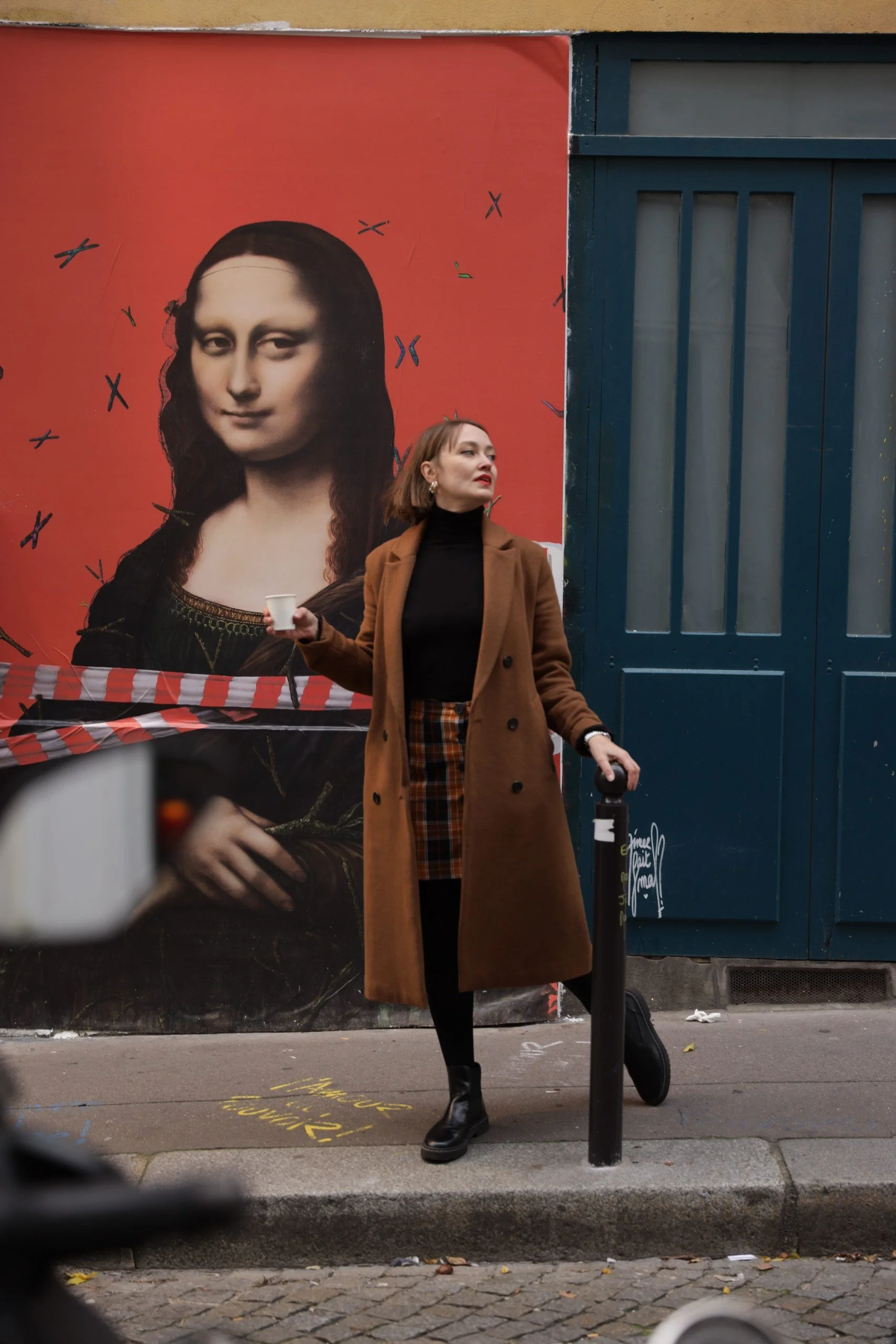 A woman walking on a city sidewalk next to a large poster of Mona Lisa on a red background. The woman is wearing a tan coat, black turtleneck, plaid skirt, black boots, and carrying a black bag and a scooter. She is holding a white paper cup.