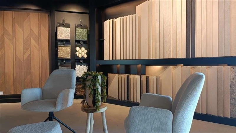 Een showroom met houten wandpanelen en decoratieve materialen, twee lichtgrijze fauteuils, een kleine tafel met een plant, lichte en donkere houten planken.