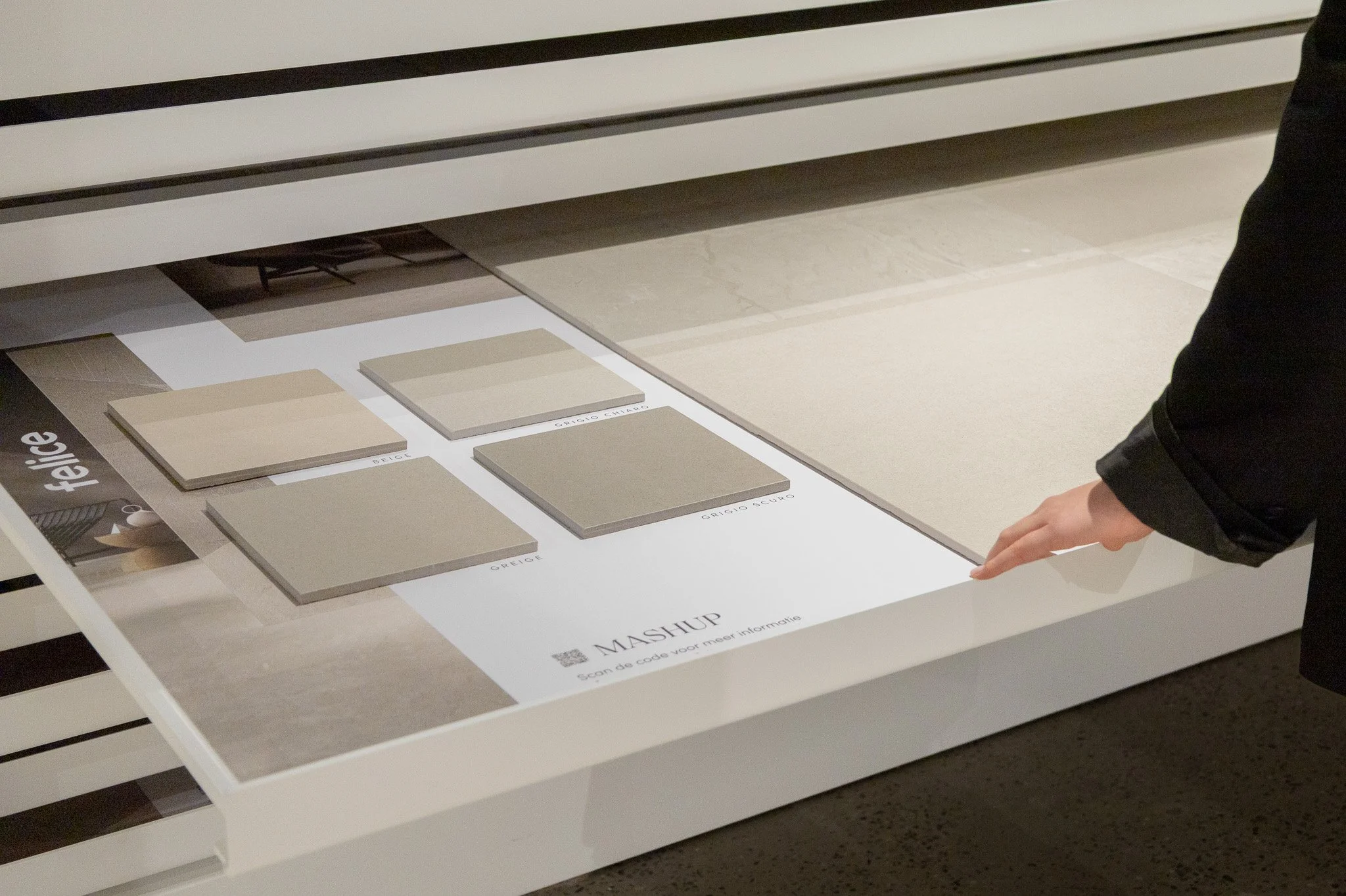 Een persoon wijst naar een display met tegels in beige, grijs en taupe tinten, met labels zoals 'sable' en 'gris chiaro', in een tegelshowroom.