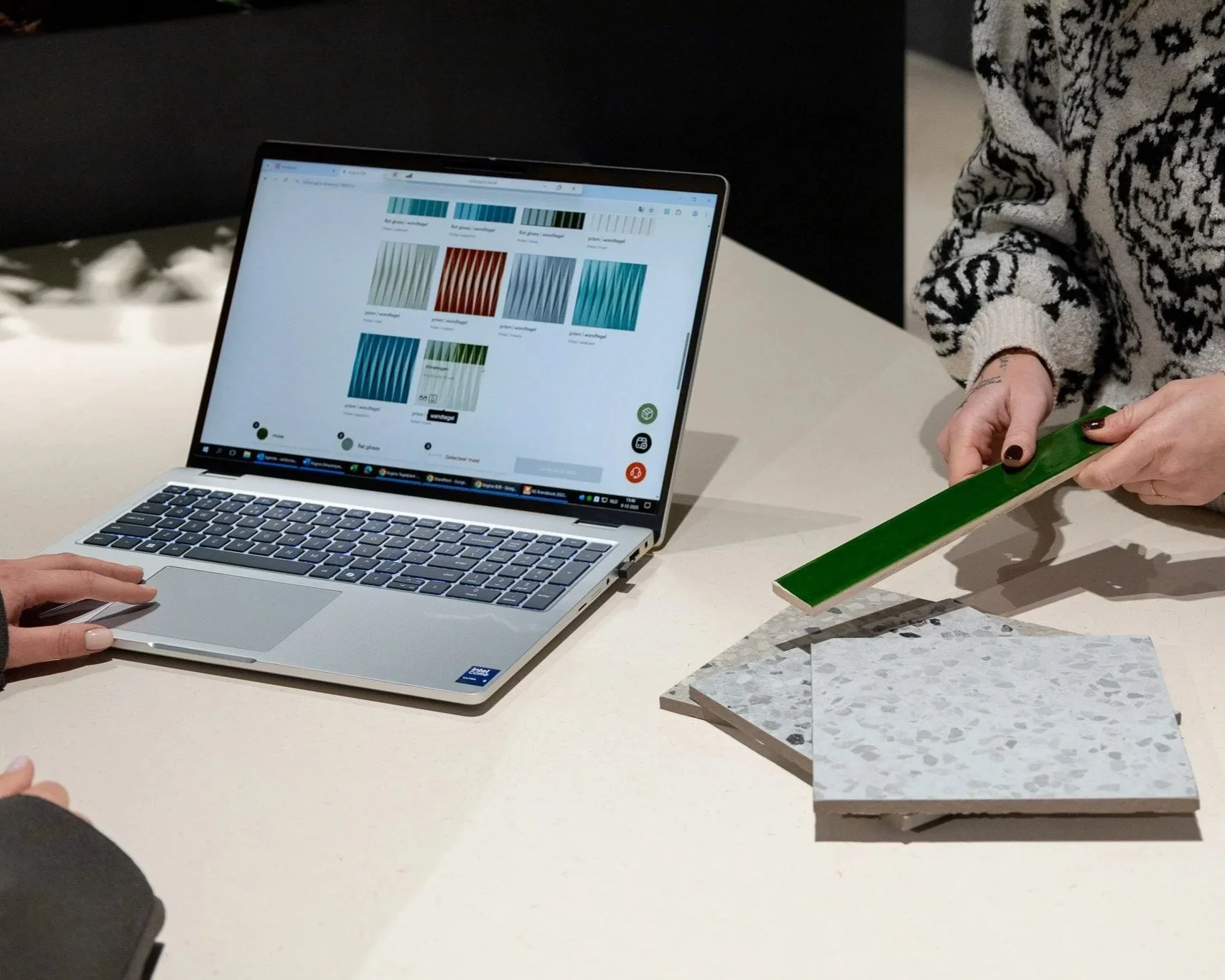 Twee mensen die kleurenstaaltjes bekijken en vergelijken, met een laptop open op de tafel.
