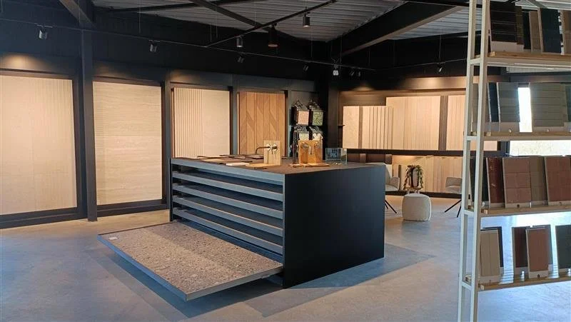 Interieur van een winkel of showroom met een zwart en houten toonbank, displayrekken met vloer- en wandpanelen, en enkele zitplaatsen in de achtergrond.