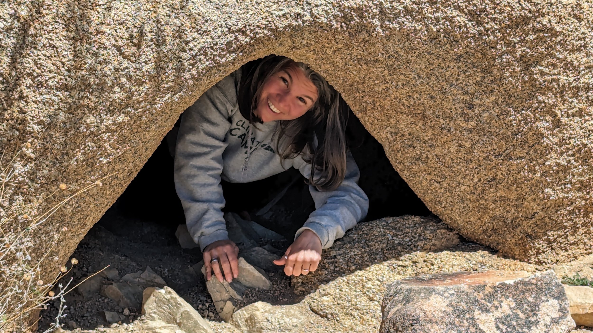 woman smiling as she peeks out from underneath a large brown boulder