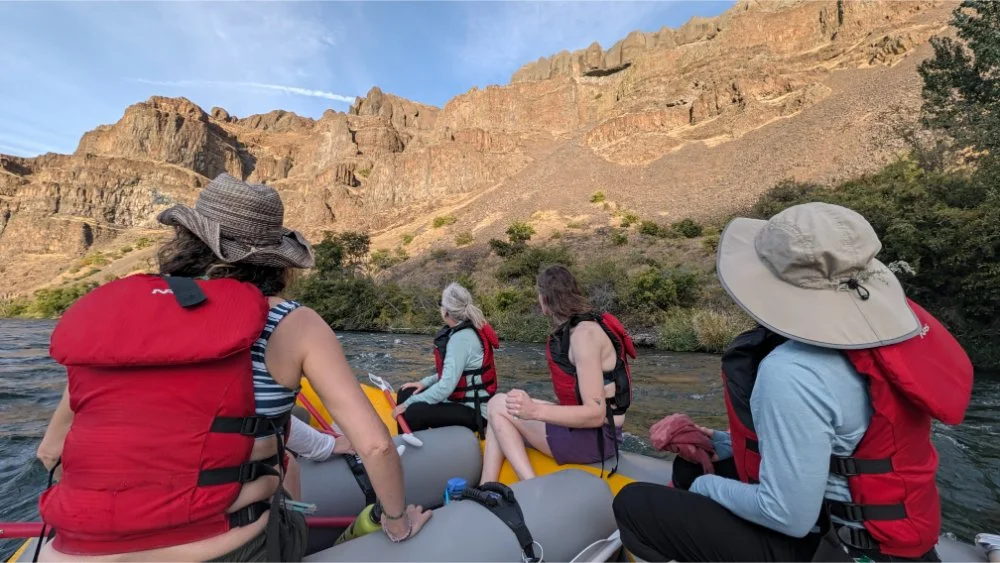 4 people in a raft on the river looking up at rocks in the sun