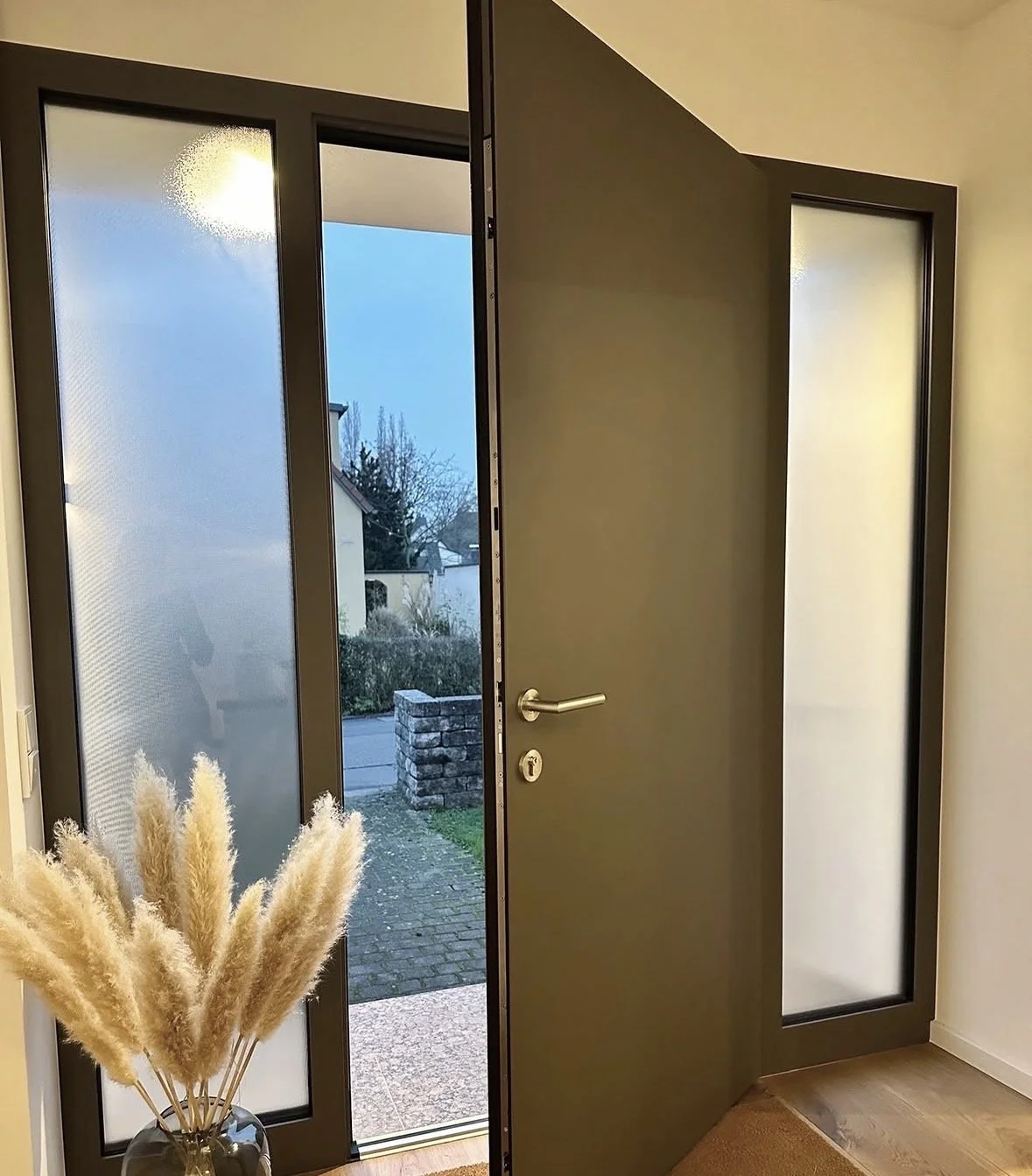 Offene Haustür mit Seitenteil, Blick nach draußen auf einen Gehweg und einen Garten, Innenraum mit Pflanze im Glasvase.
