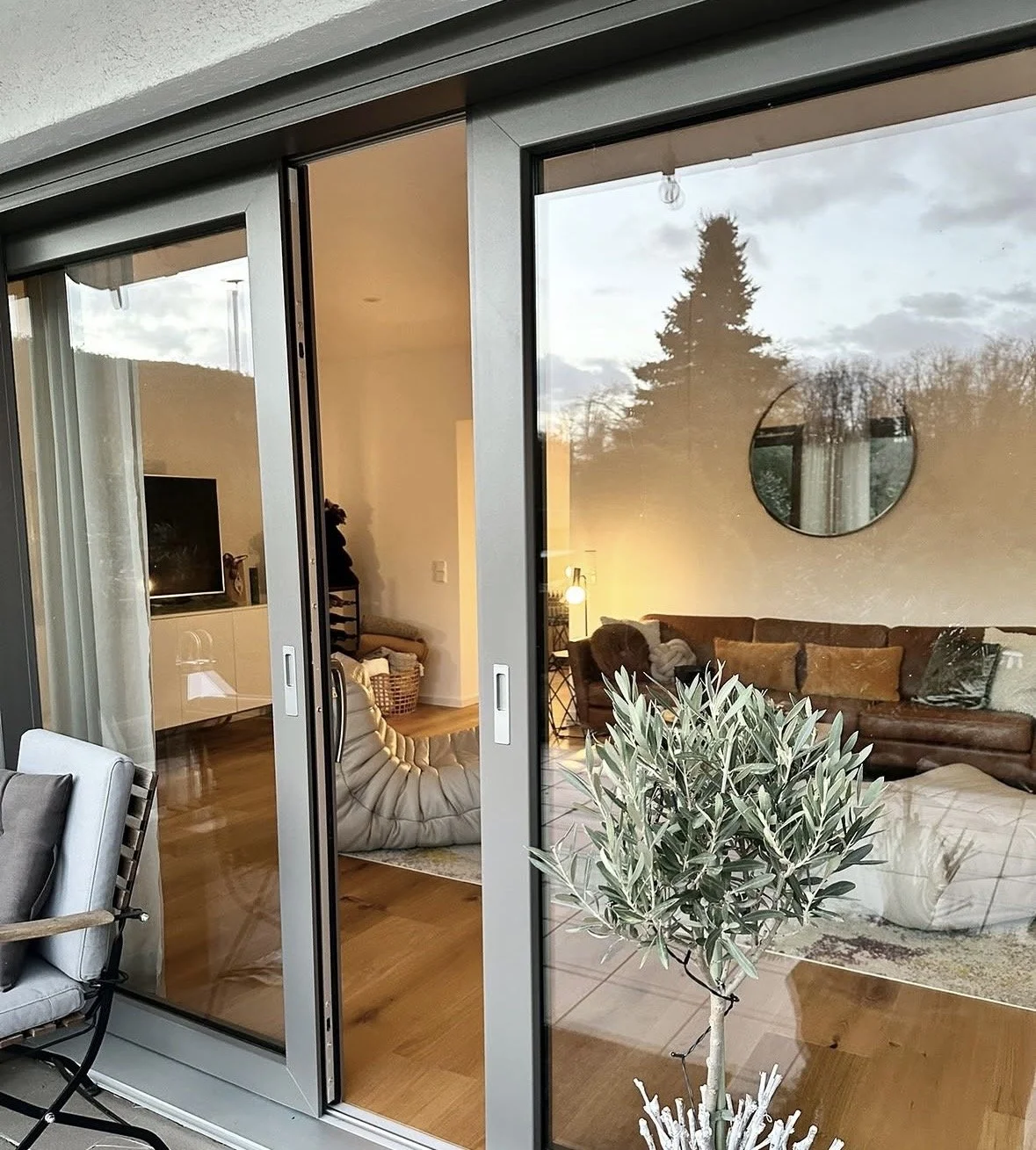 Ein Blick durch eine große Glasschiebetür in ein Wohnzimmer mit Sofa, Pflanzen und einem Fernseher, bei Dämmerlicht im Freien