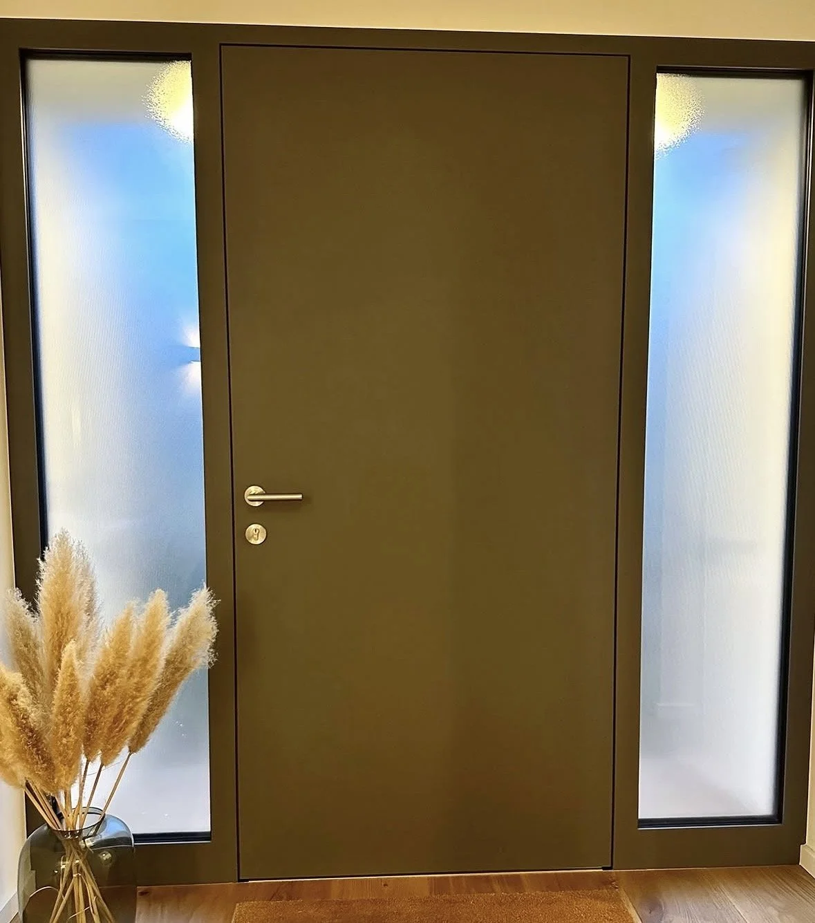 Moderne Eingangstür in Dunkelgrün mit seitlichen gläsernen Seitenteilen, neben einem schwarzen Keramikvase mit Pampasgras, auf Holzboden.