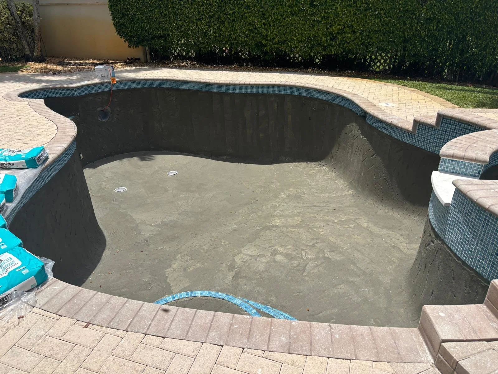 Empty swimming pool under construction, with stone-paved deck and green bushes in the background. Pool resurfacing and renovation project completed by Lux Pools