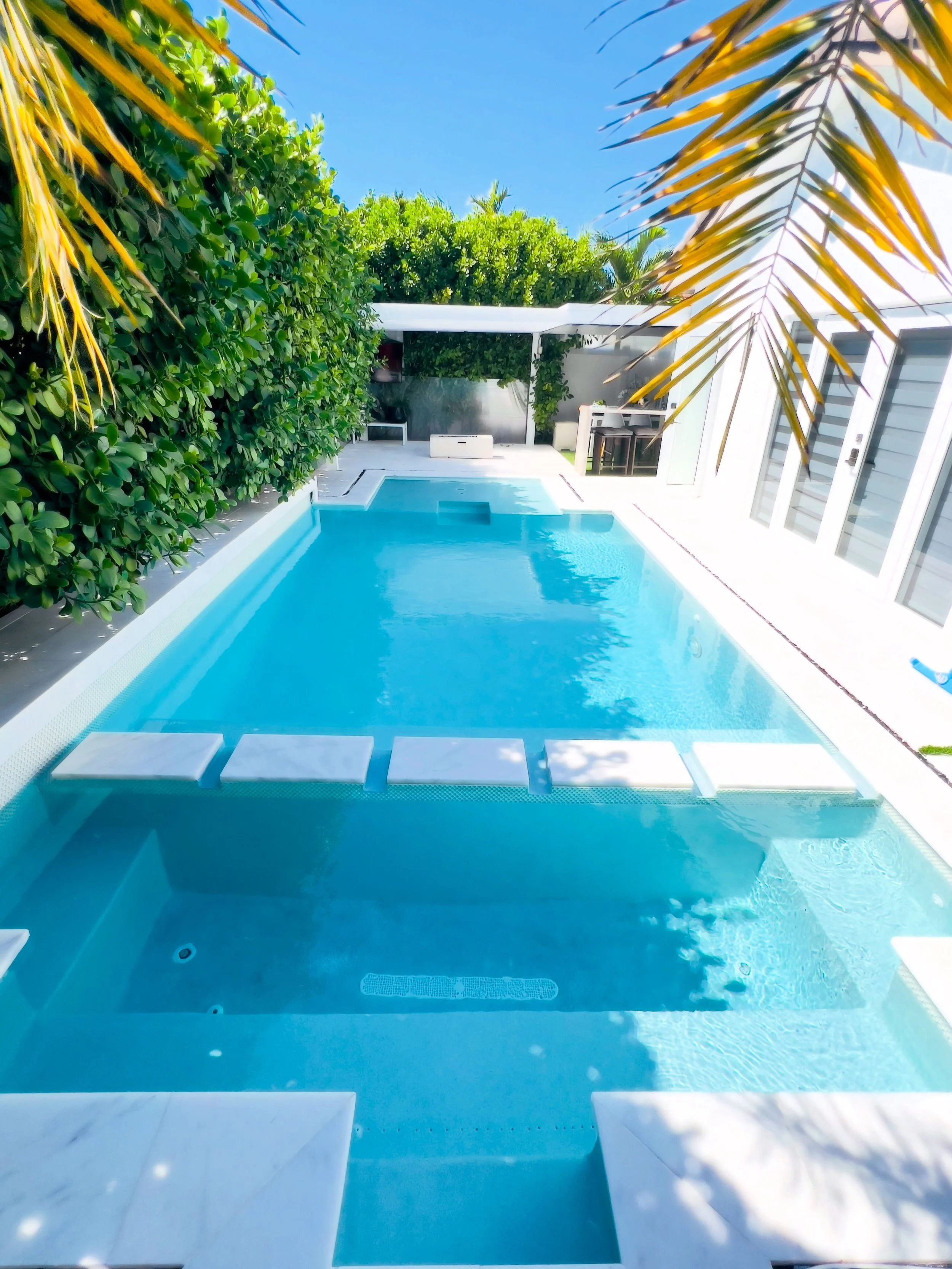 A modern backyard swimming pool surrounded by lush green hedges and palm trees, with clear blue sky overhead.