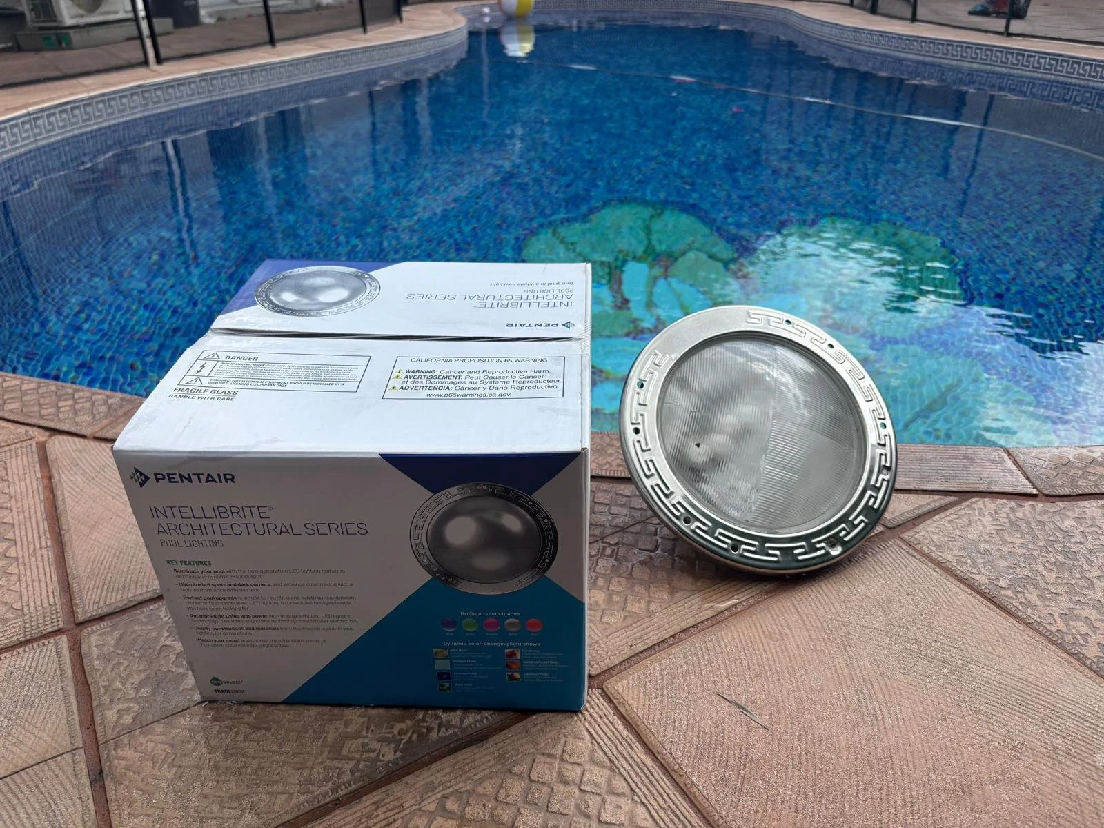 A box of Pentair IntelliBrite architectural pool lighting and a pool light fixture placed on a brick pool deck with a swimming pool in the background. Swimming pool lighting installation in Miami-Dade County