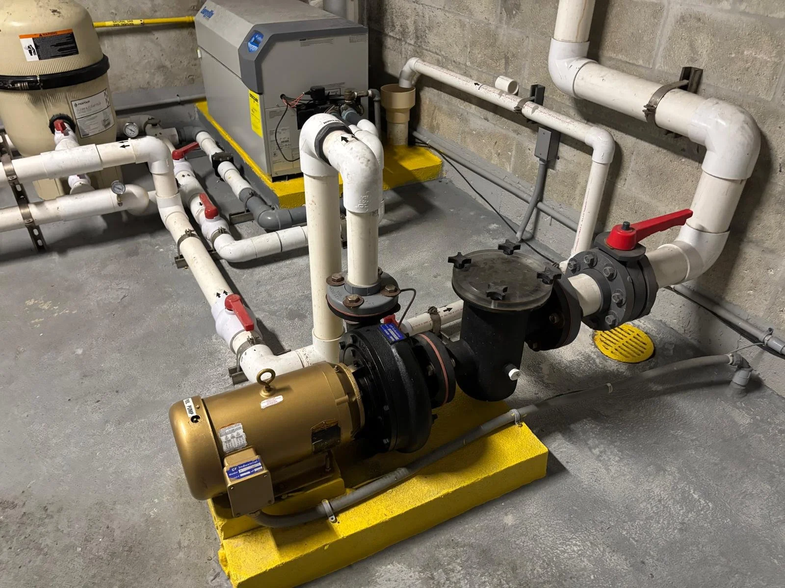 Industrial pump system with white piping, a large black pump, a gold motor, and a gray box on a yellow platform in a basement setting. Pool equipment repair and installation service