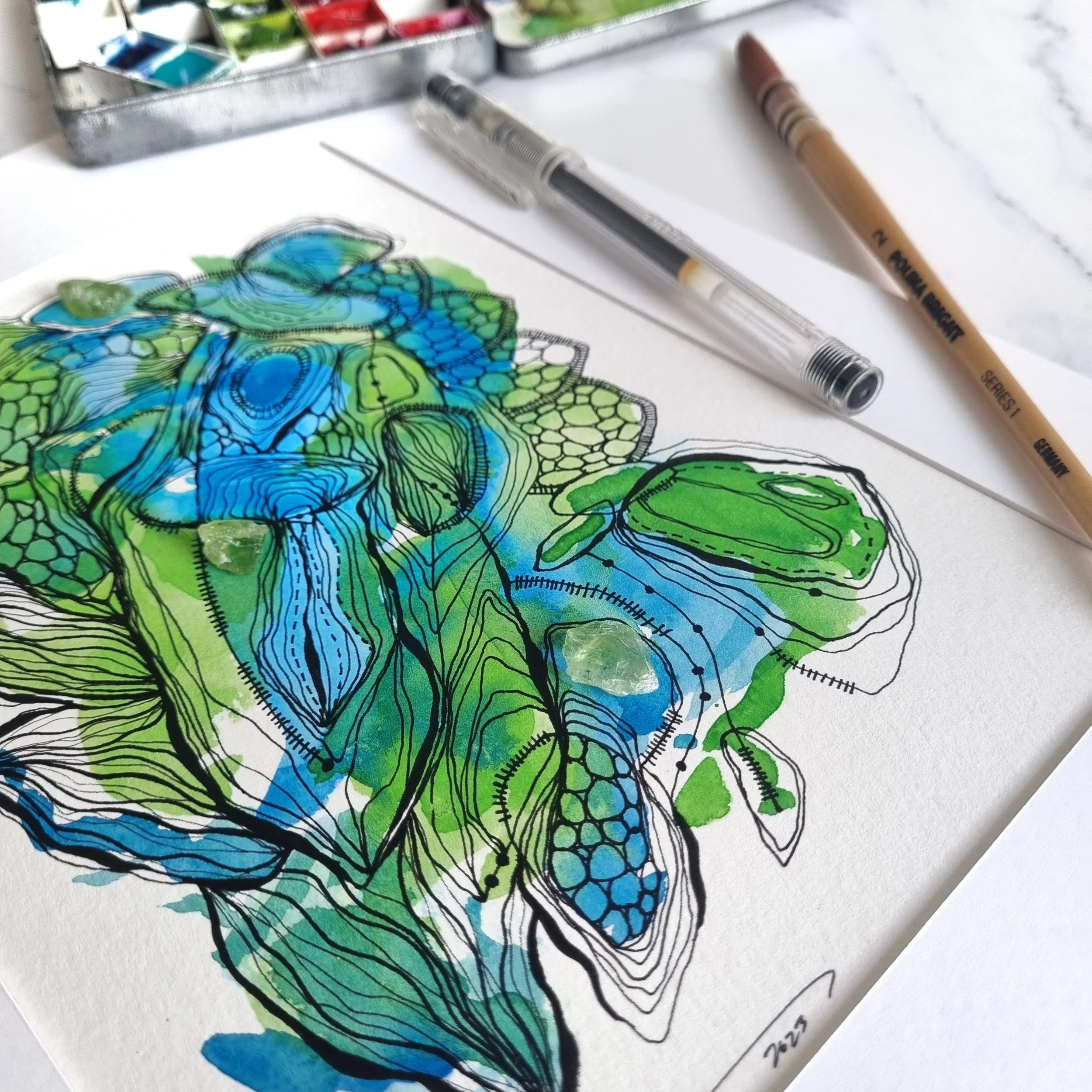 Colorful abstract watercolor painting of organic shapes with black ink outlines, placed on white paper with art supplies nearby.