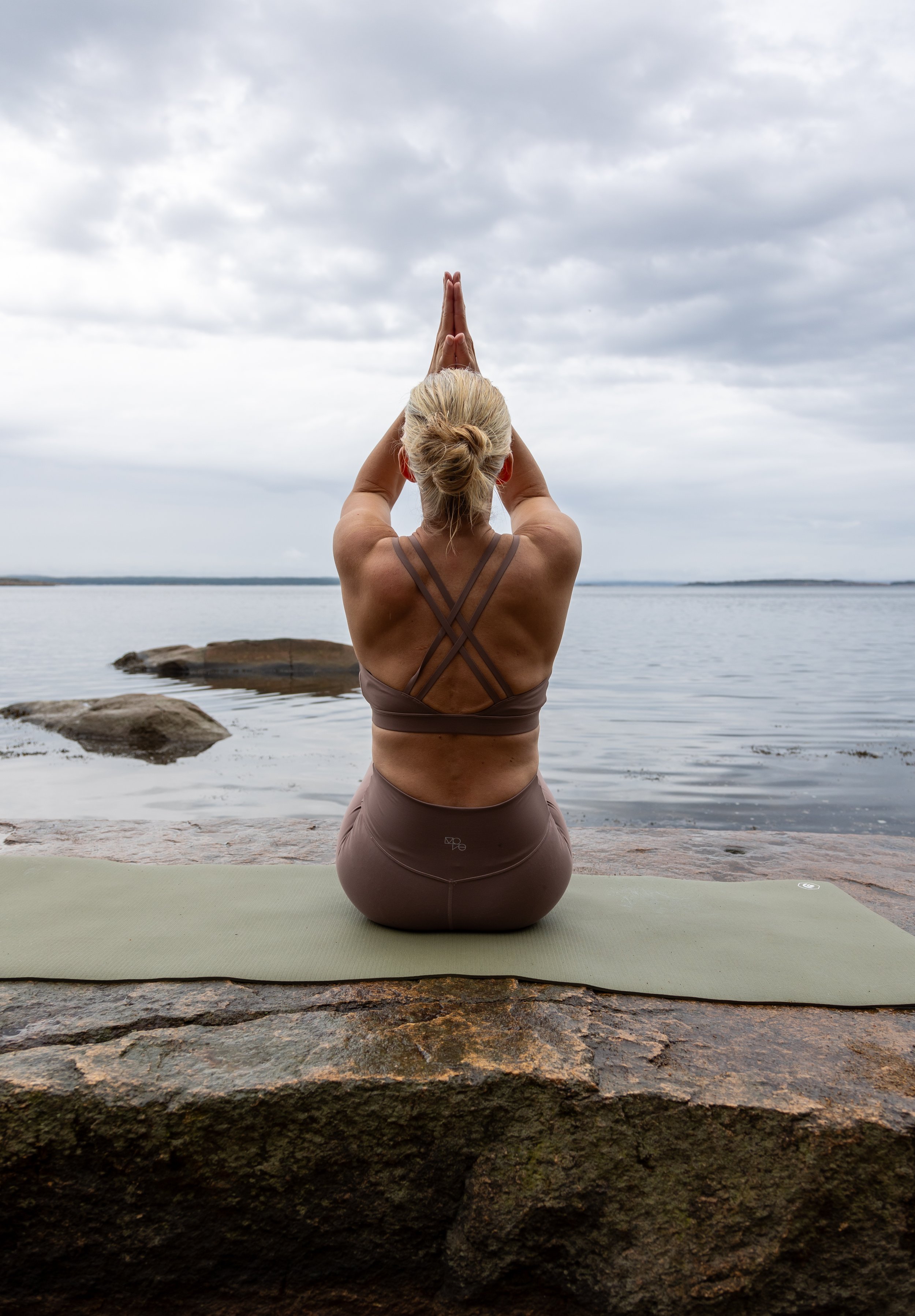 Person som gjør yoga ved sjøen, sittende på en yogamatte med hendene løftet over hodet i meditasjonsstilling, under overskyet himmel.