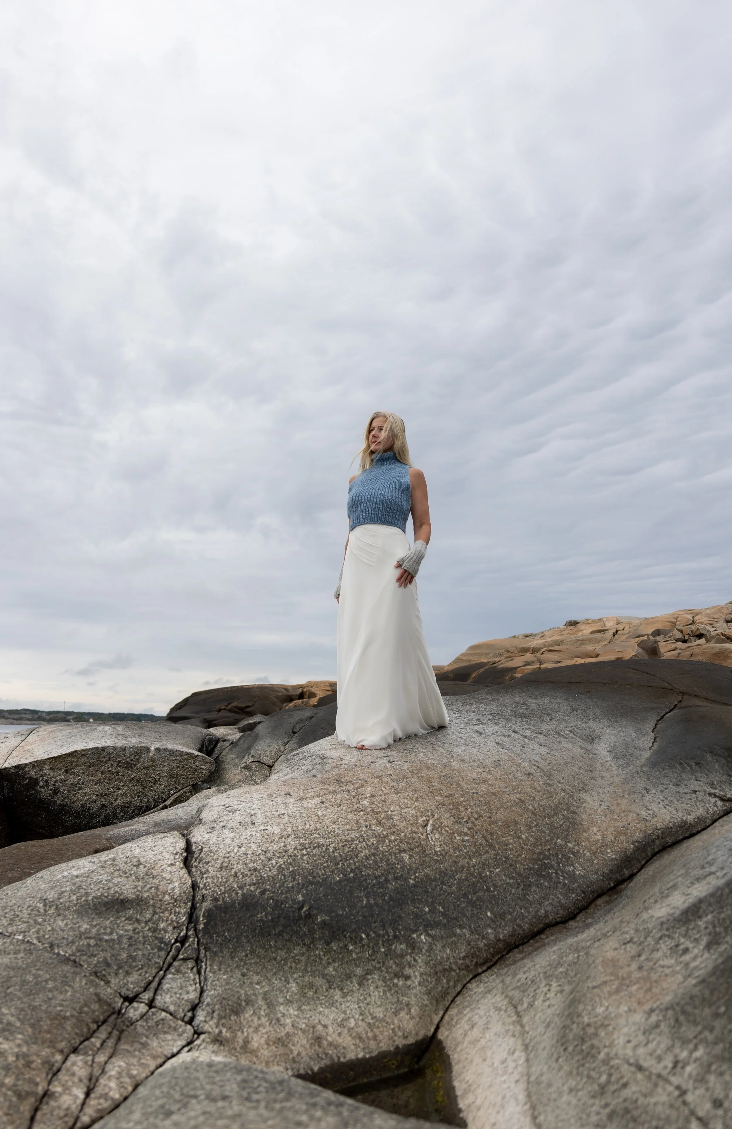 En kvinne i blå genser og hvit skjørt står på en stein ved kysten under en overskyet himmel.