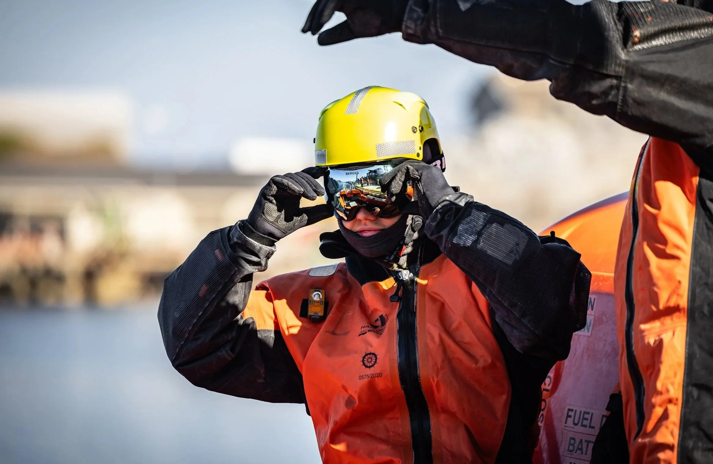 En person i oransje og svart redningsdrakt og gul hjelm tar på seg vernebriller, med en annen person ved siden av seg.