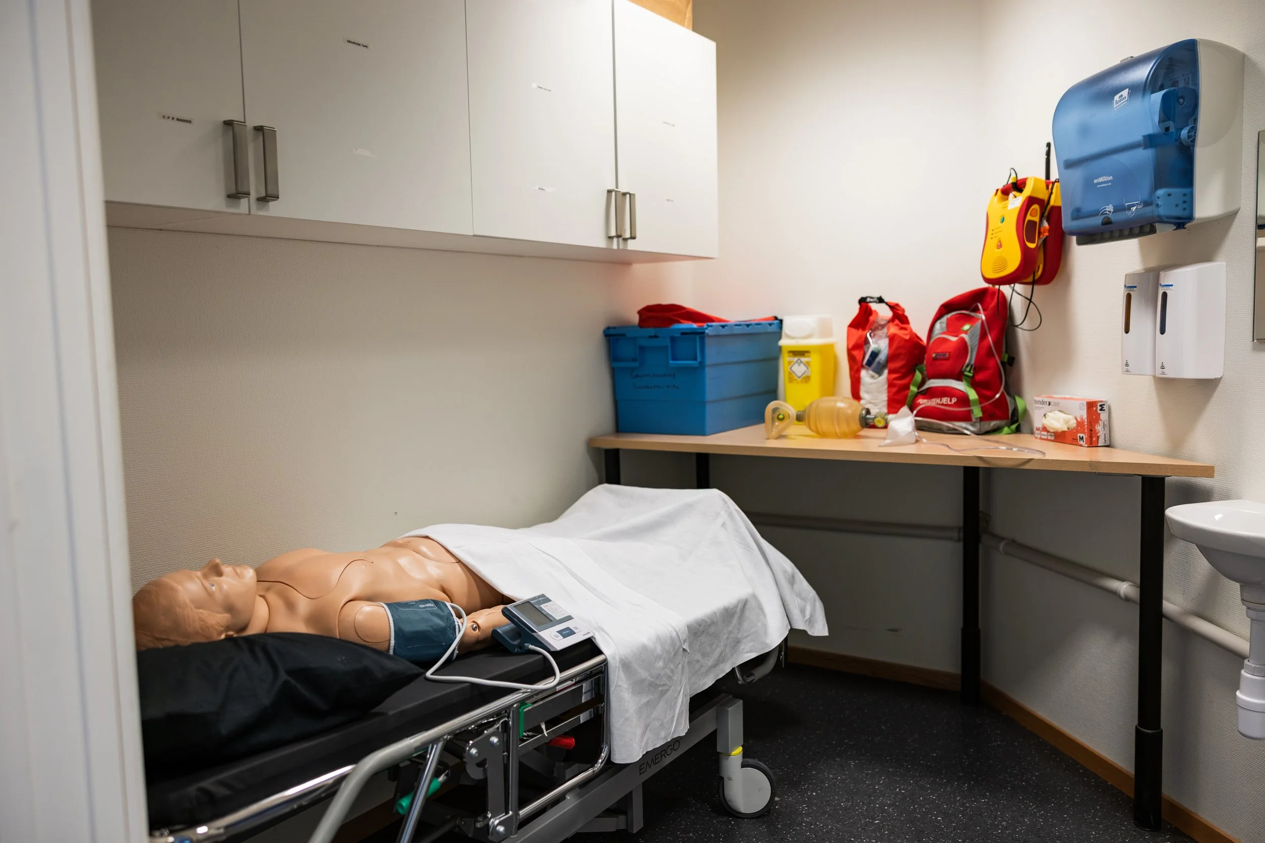 En redningsdukke på senga i et treningsrom med medisinsk utstyr og medisiner i bakgrunnen.